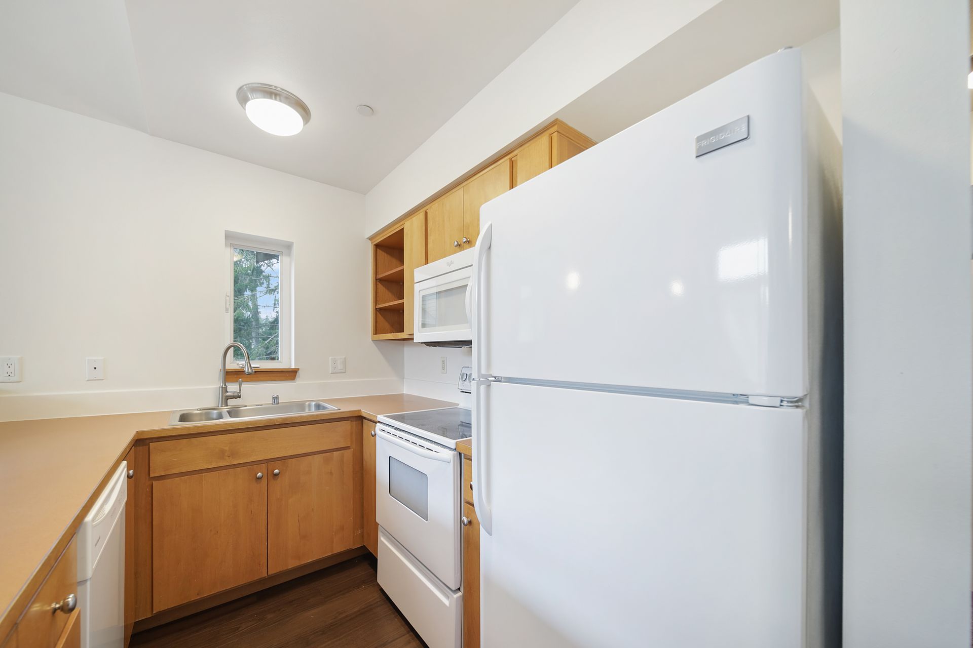 Bright kitchen with white appliances, wood cabinets, and a sink by a small window.