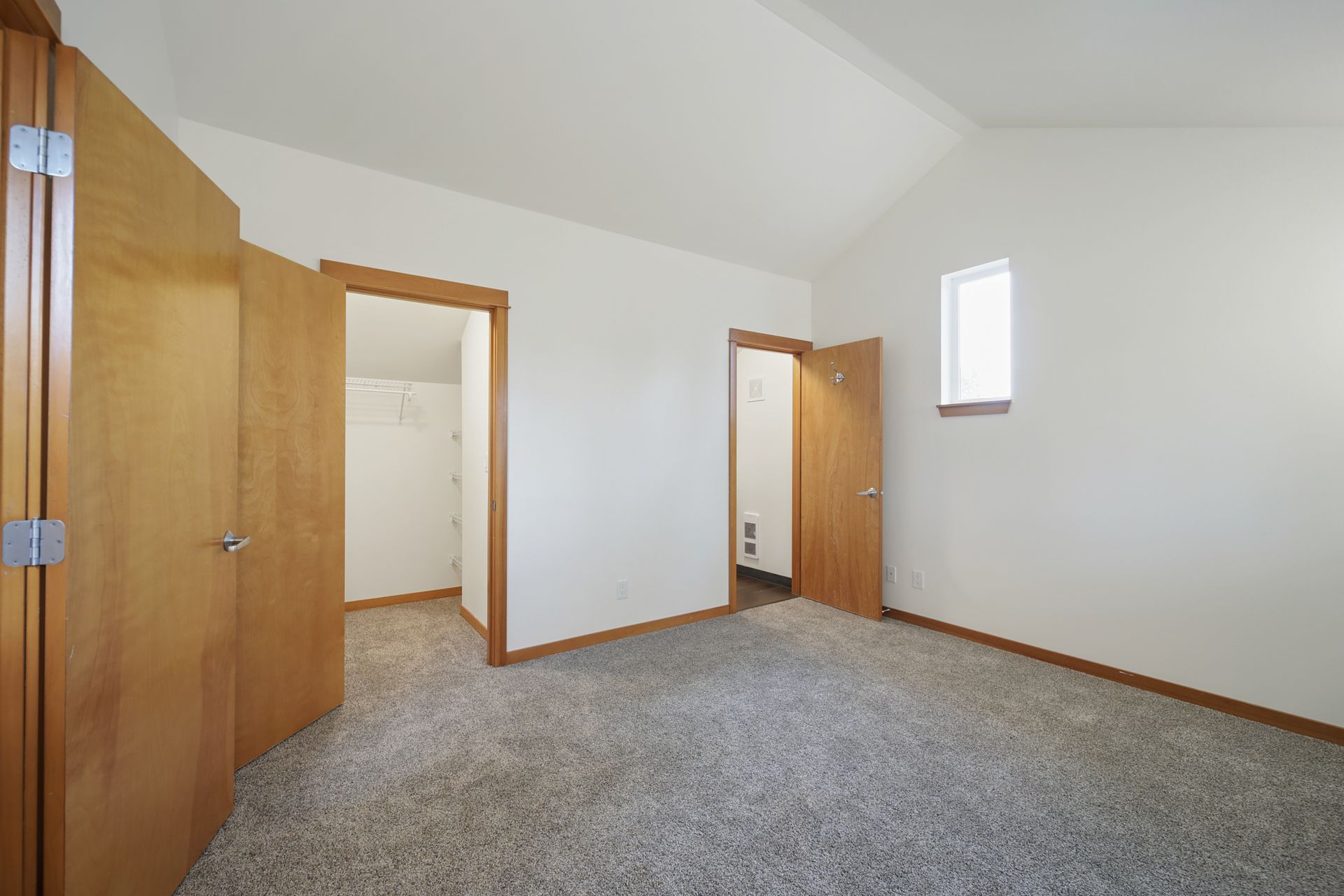 Empty carpeted bedroom with white walls, wooden trim, open closet doors, and a small high window