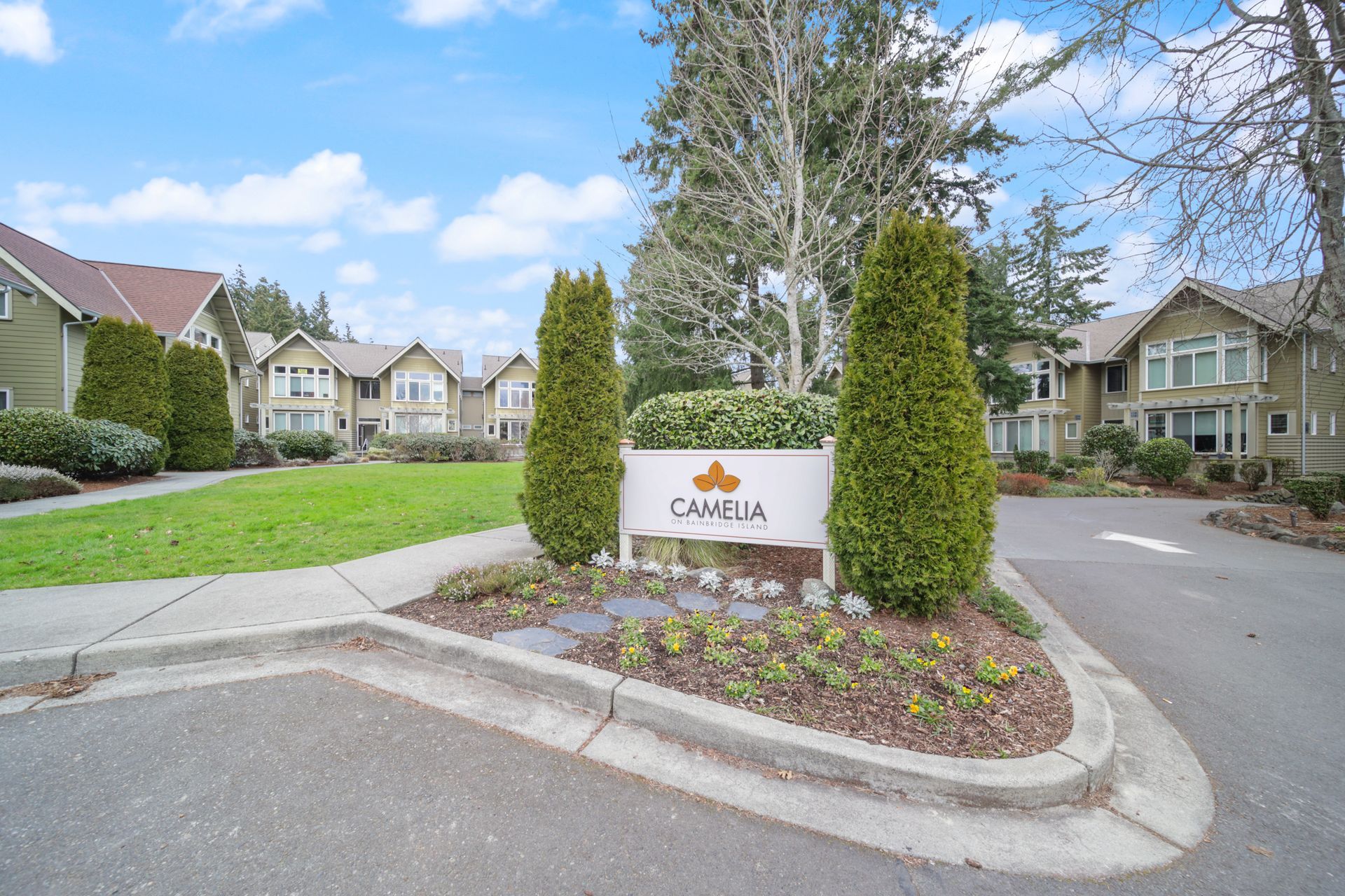 Apartment complex entrance with a stone sign, trimmed shrubs, and townhouses under a blue sky