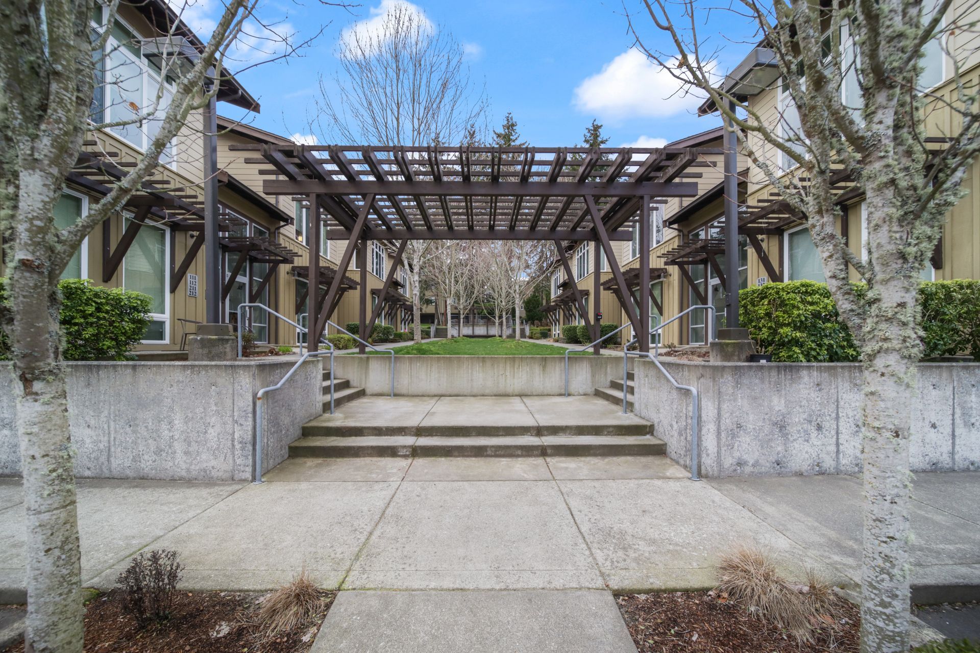 Modern courtyard entrance with concrete steps, wooden pergola, and leafless trees under a cloudy sky