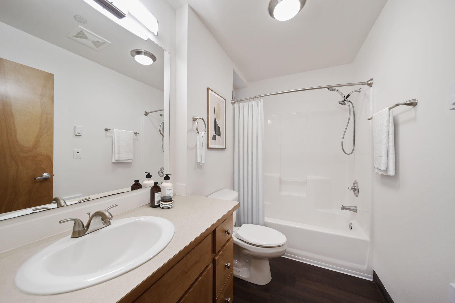 Bright bathroom with vanity sink, mirror, toilet, and white shower/tub combo with dark wood floor.
