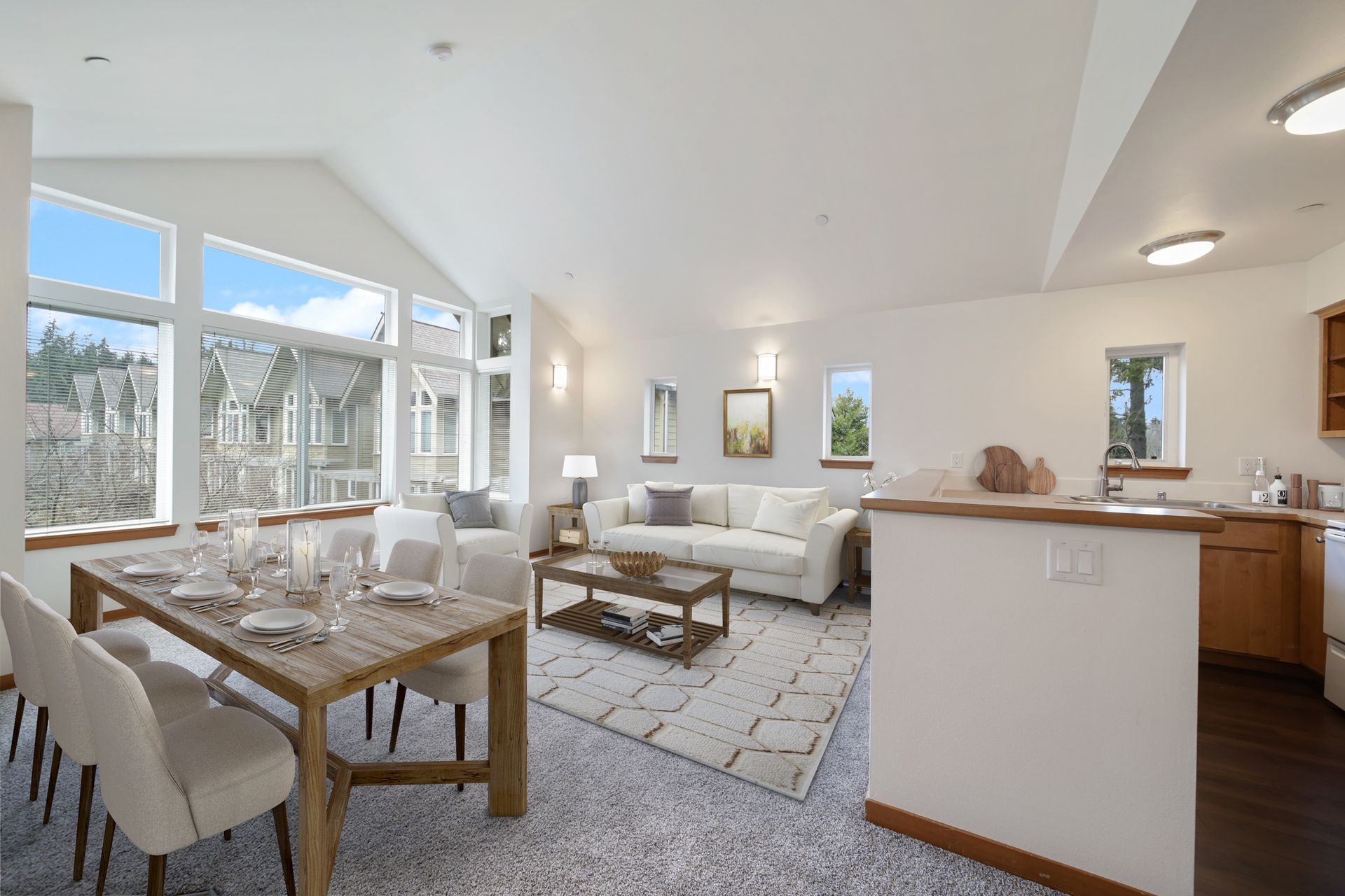 Bright open-plan living and dining room with white sofas, wood table, and large windows overlooking a patio
