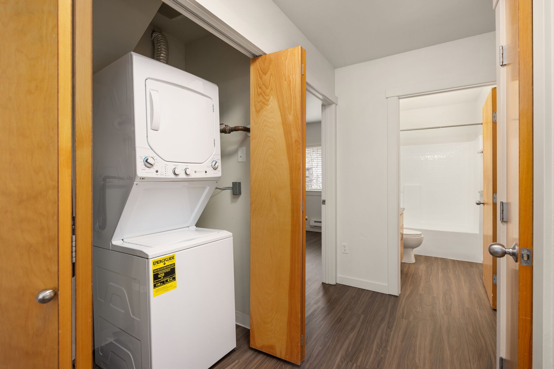 Laundry closet with stacked washer and dryer beside an open apartment hallway and bathroom.