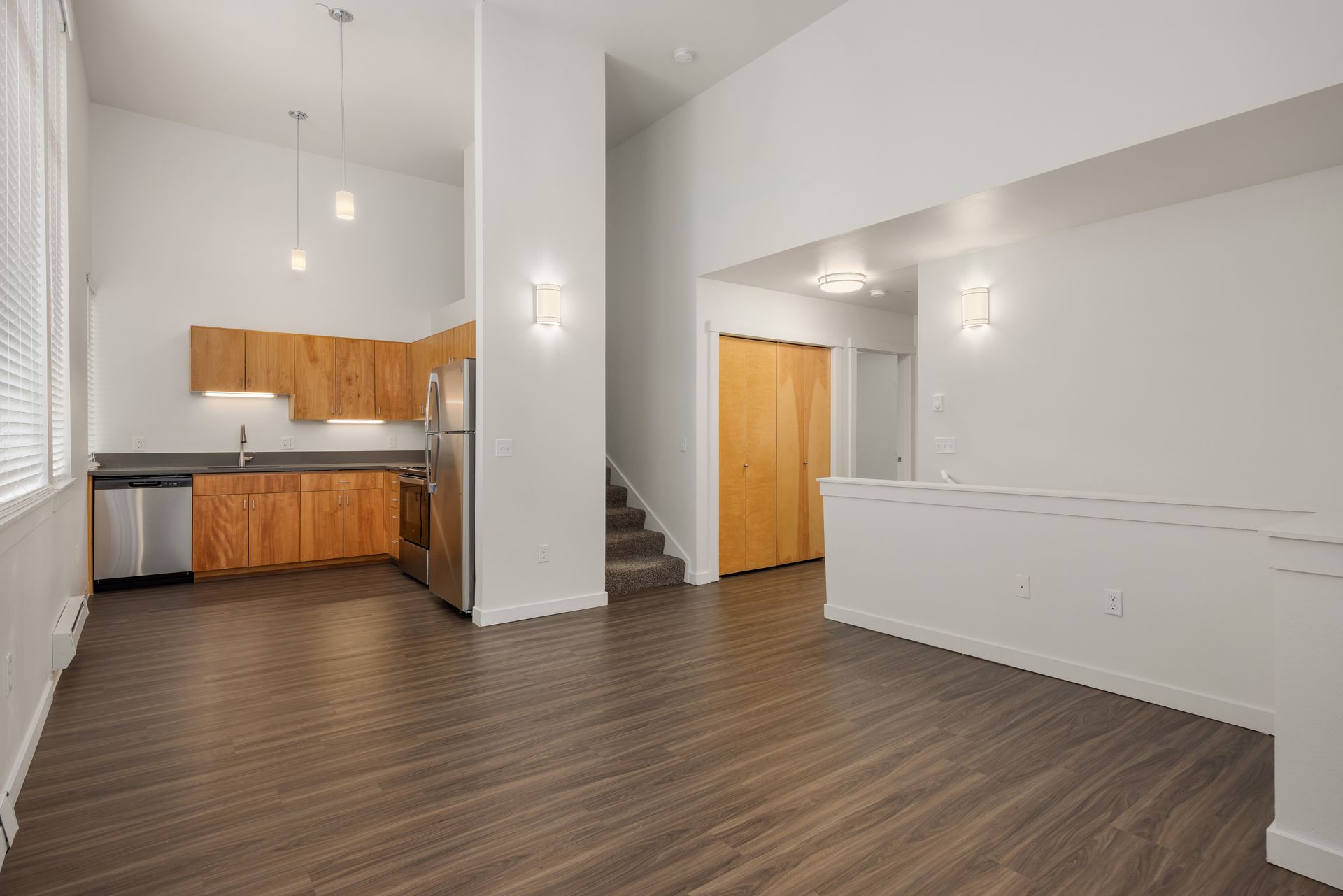 Spacious empty loft with wood floors, white walls, kitchen area, and staircase to an upper level