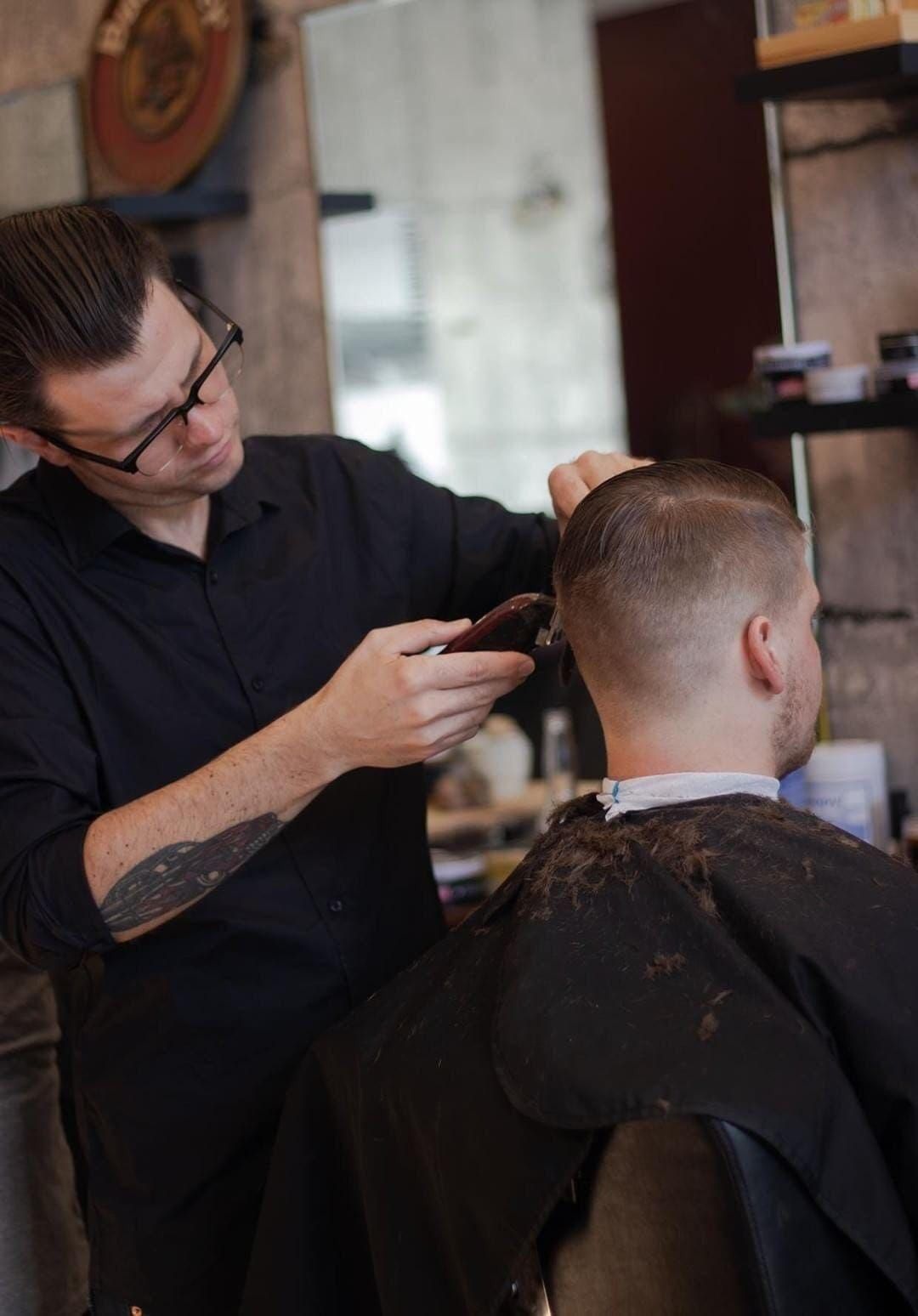 Barber cutting a man's hair with clippers. Both are in a barber shop. The barber wears glasses and a black shirt. — Eagle Tamer Barbershop in Stratford, QLD