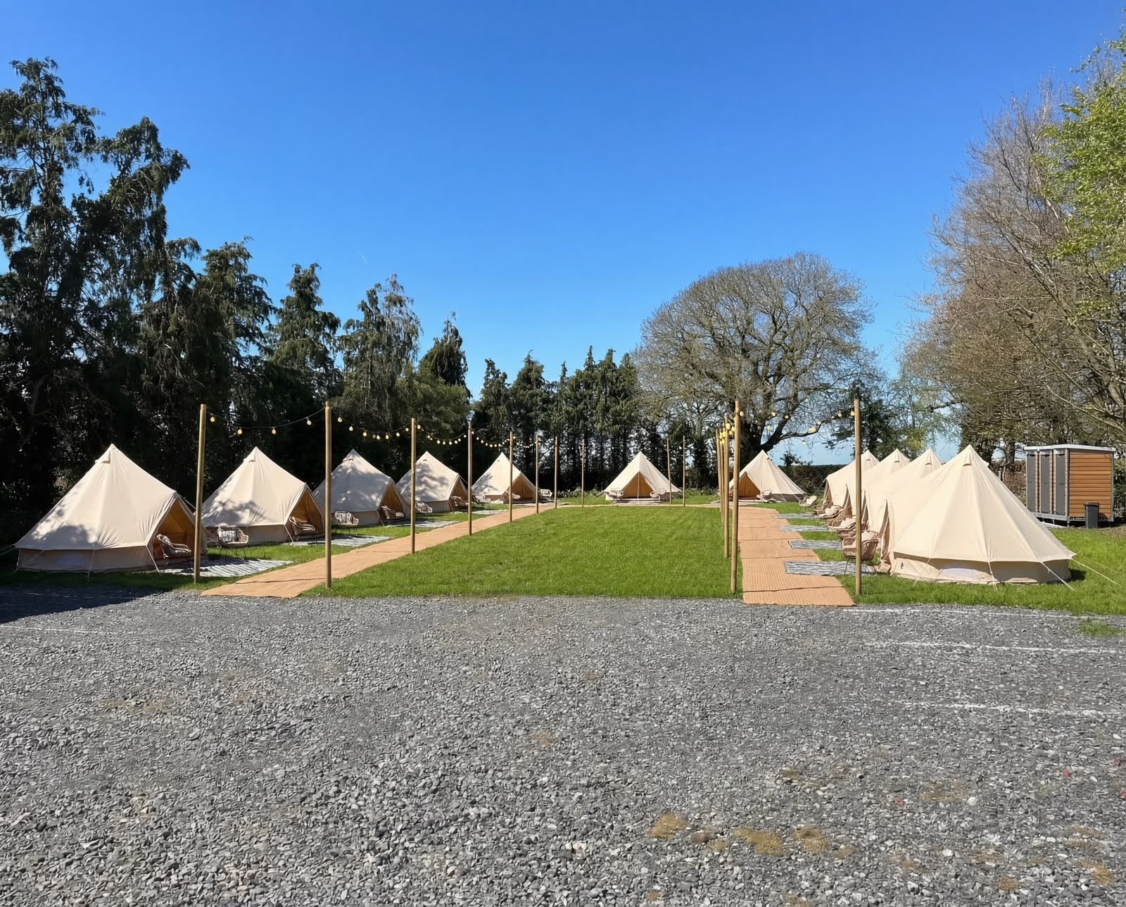 Row of beige canvas tents on grass beside gravel, under a clear blue sky