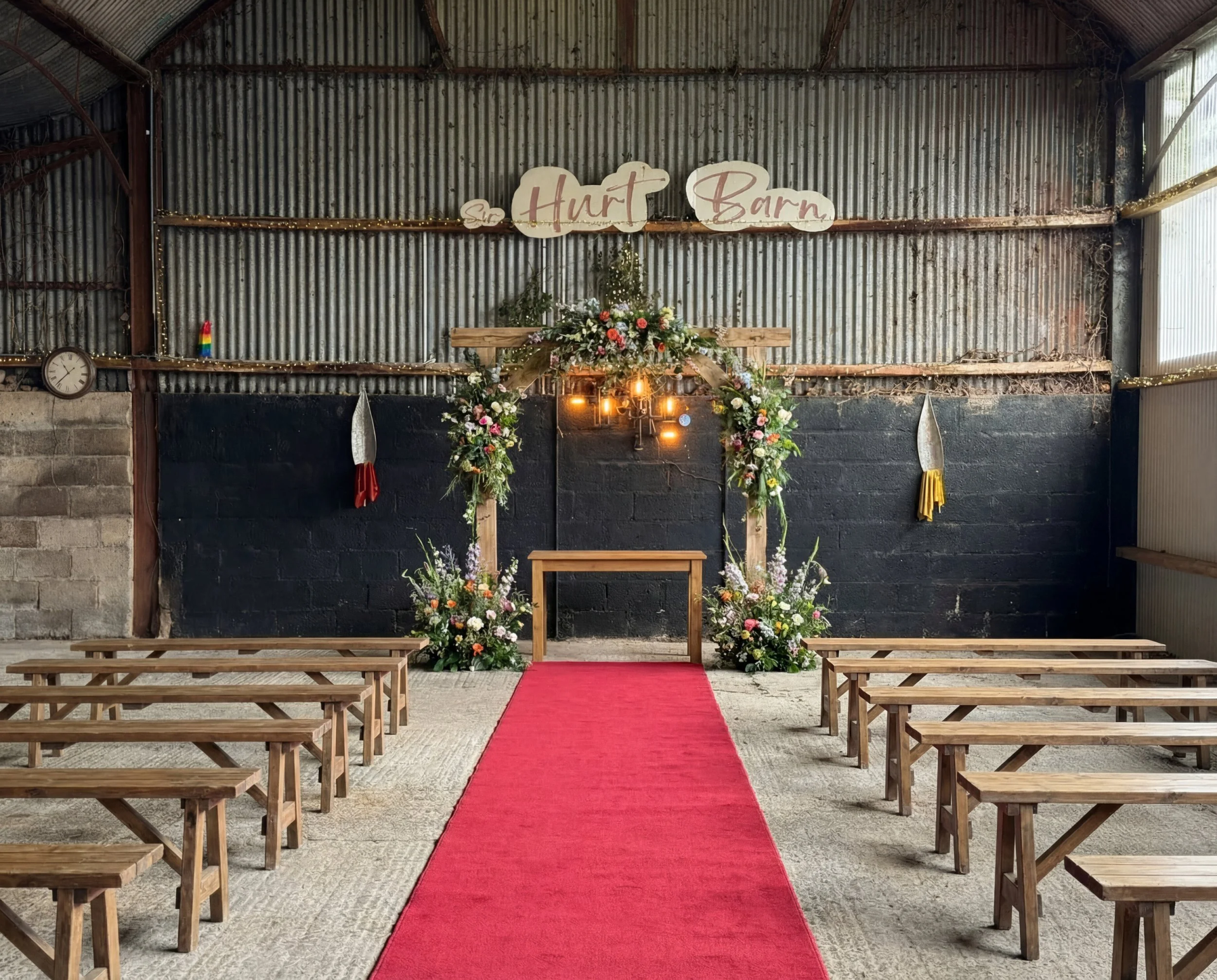 Rustic indoor wedding aisle with red carpet, wooden benches, and floral arch under corrugated metal walls
