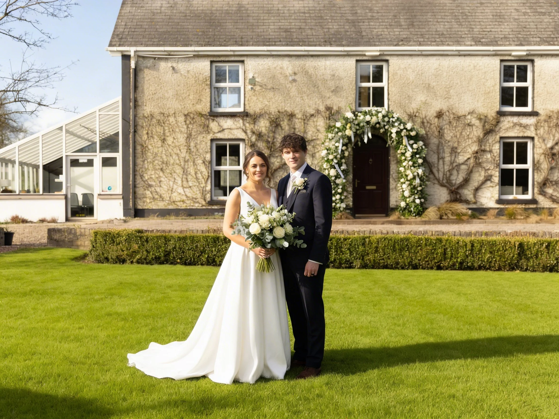 Bride and groom posing on a lawn in front of a stone house and floral arch
