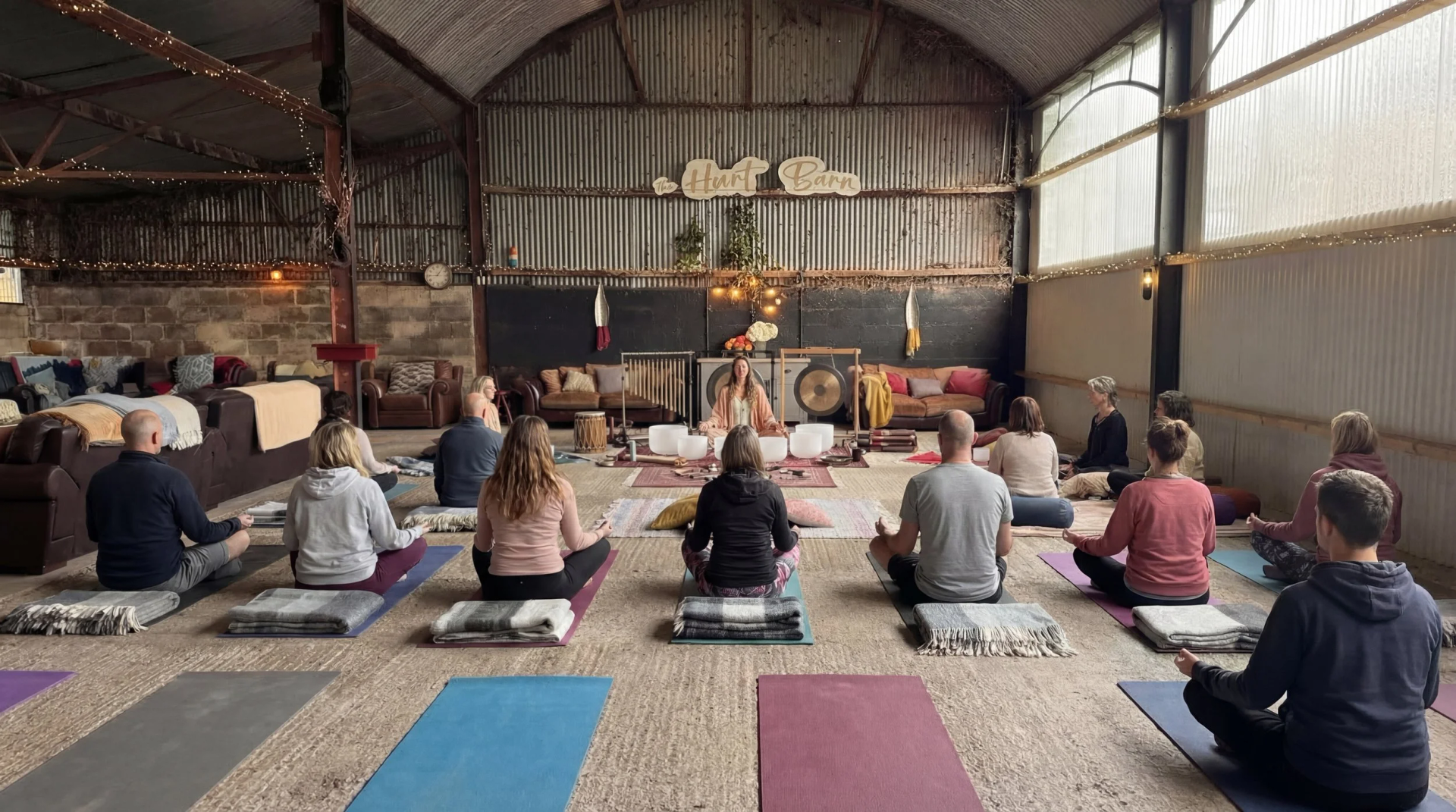 People seated on yoga mats in a rustic hall, facing a lit altar with candles and instruments.
