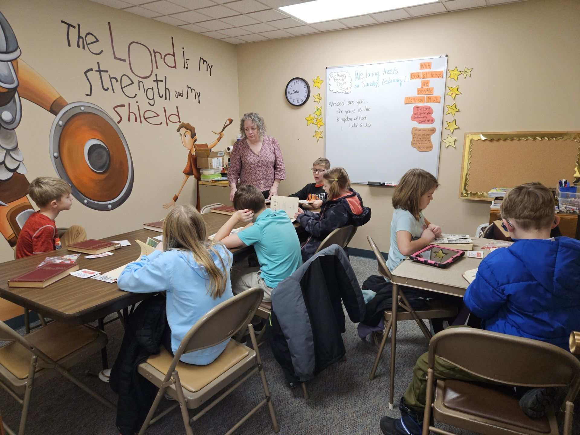 Students sit at desks in a classroom, with a teacher at the front near a whiteboard and wall mural.