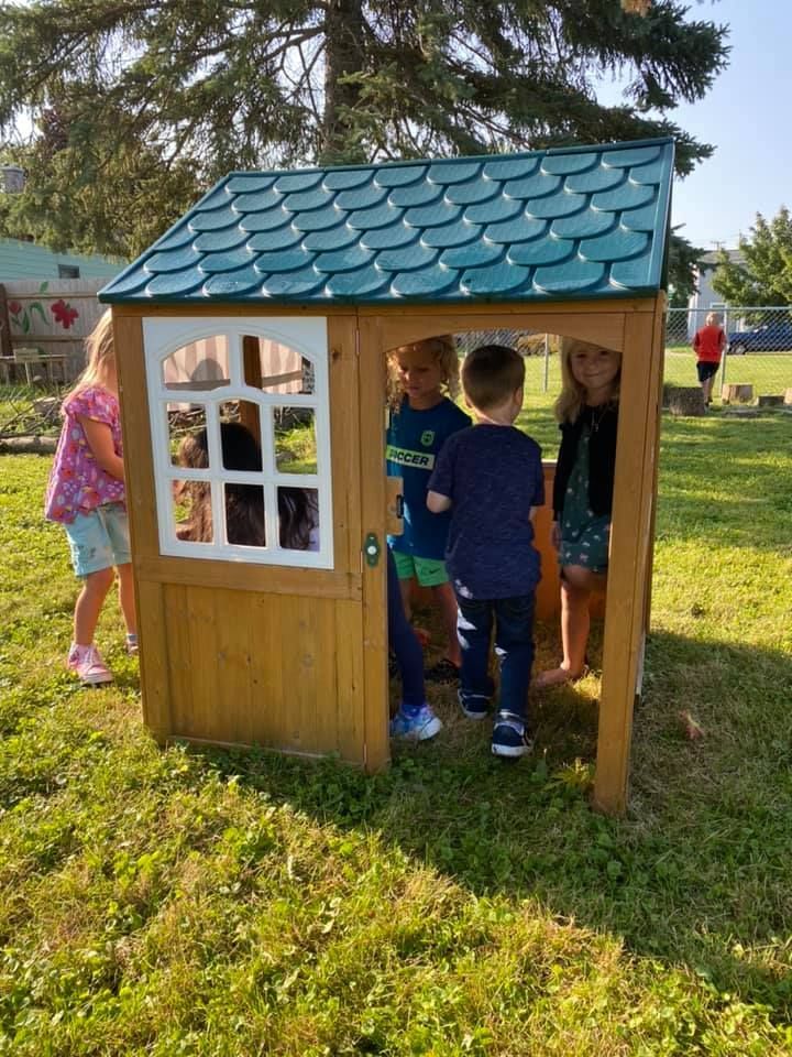 Children playing inside a small wooden playhouse with a blue roof on a sunny grassy lawn.
