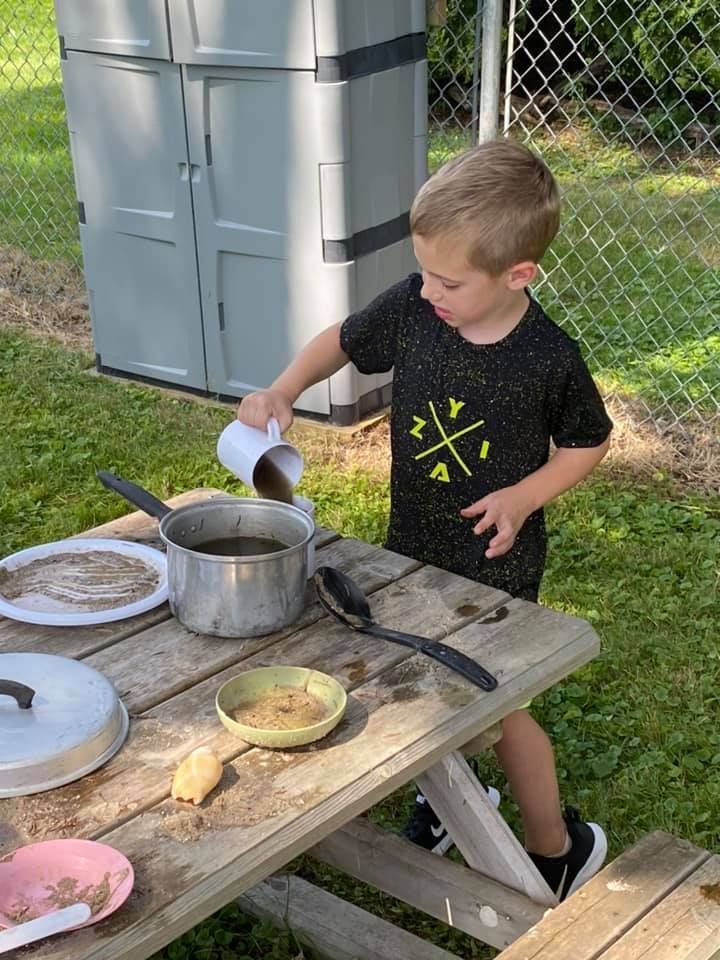 Child pouring liquid into a pot at an outdoor table in a backyard, with dishes and cookware nearby
