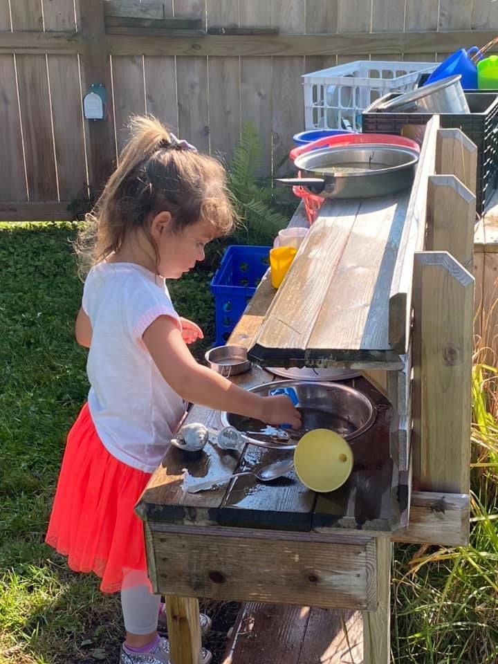 Child playing at an outdoor toy kitchen by a wooden fence, wearing a white shirt and red skirt.