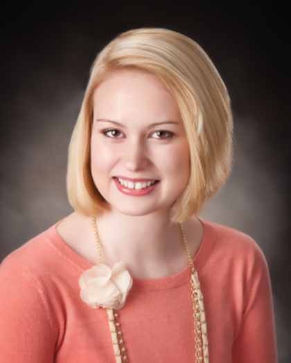 Smiling blonde portrait against a dark background, wearing a coral top and long necklace