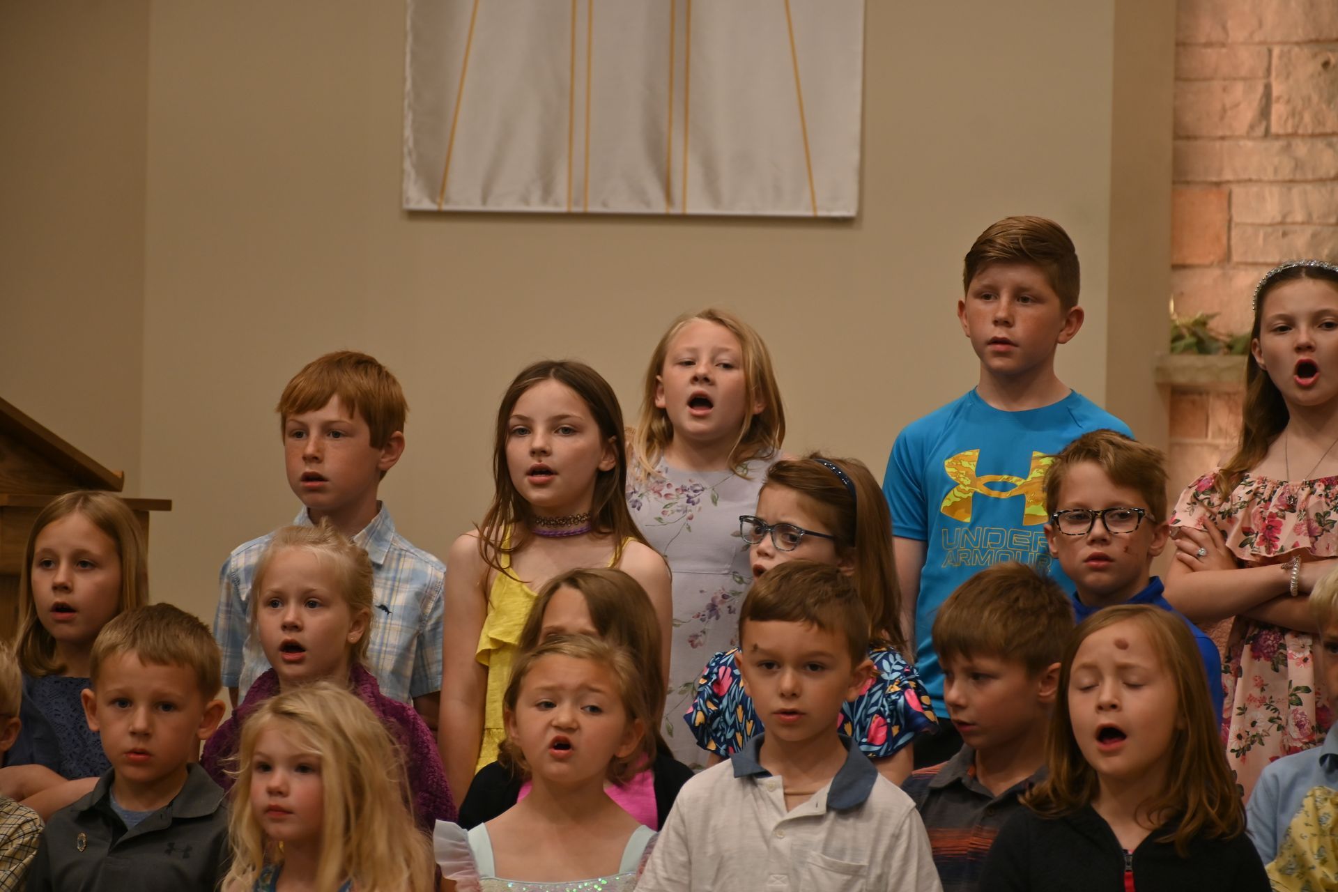 Children singing in a choir, with several open-mouthed mid-song on a church stage