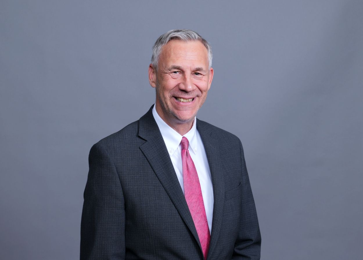 Smiling man in a dark suit with a pink tie against a gray studio backdrop