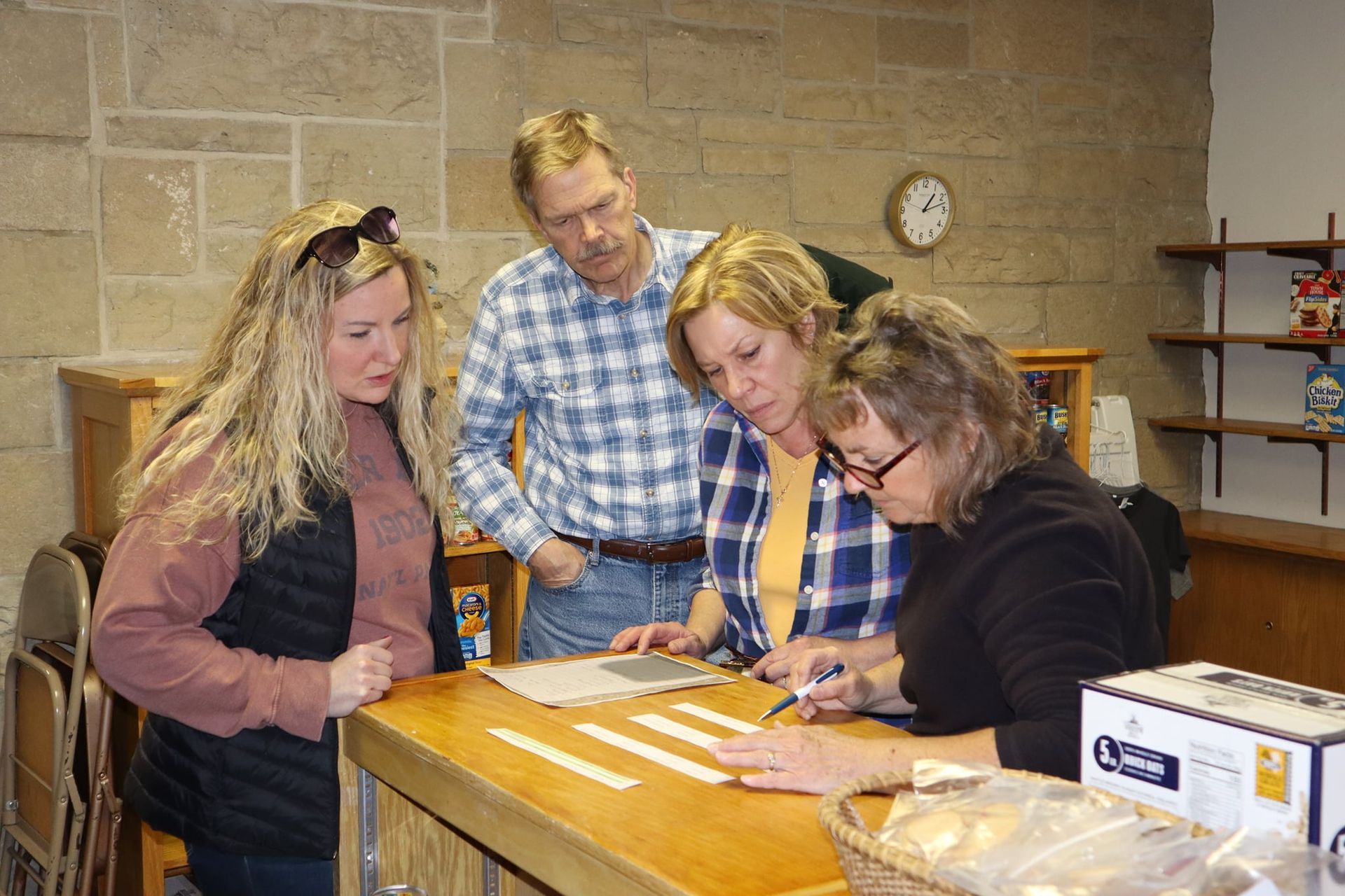 Four people lean over papers at a counter in a kitchen or classroom setting