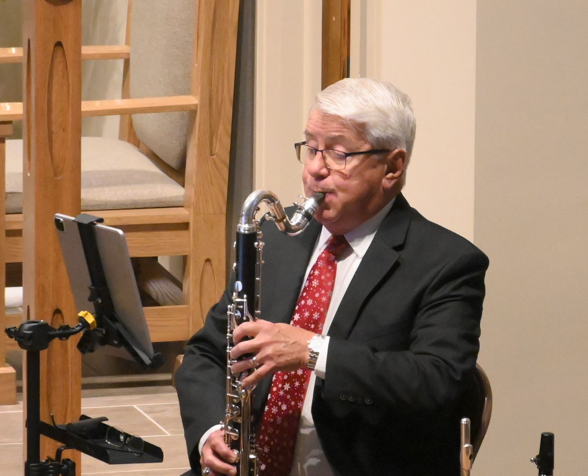Musician playing a clarinet onstage, wearing a black suit and red tie, beside a wooden ladder backdrop