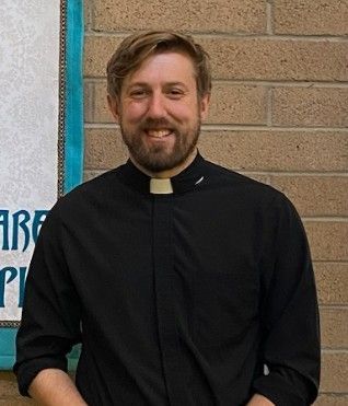 Smiling priest in a black clerical shirt with white collar standing against a brick wall.
