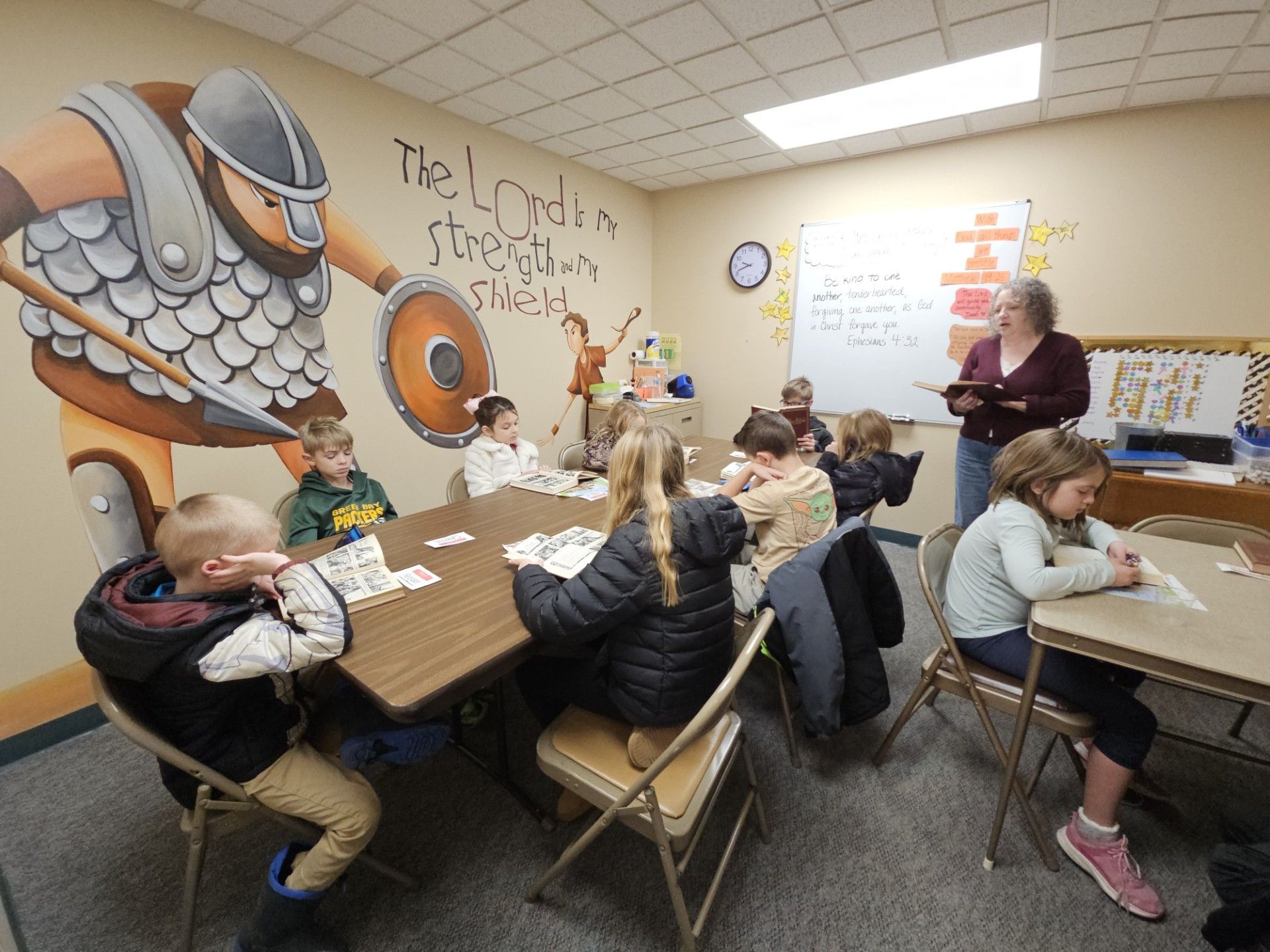 Children seated at a classroom table during a lesson, with a teacher standing near the front wall.