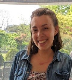 Smiling person in a denim shirt standing indoors by a window with trees outside.