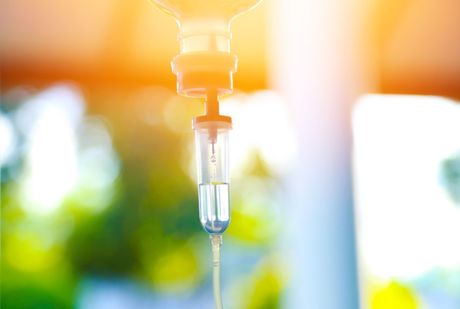 Nahaufnahme eines intravenösen (IV) Tropfes mit Fokus auf die Tropfkammer, mit einer unscharfen, hell erleuchteten Umgebung im Hintergrund.
