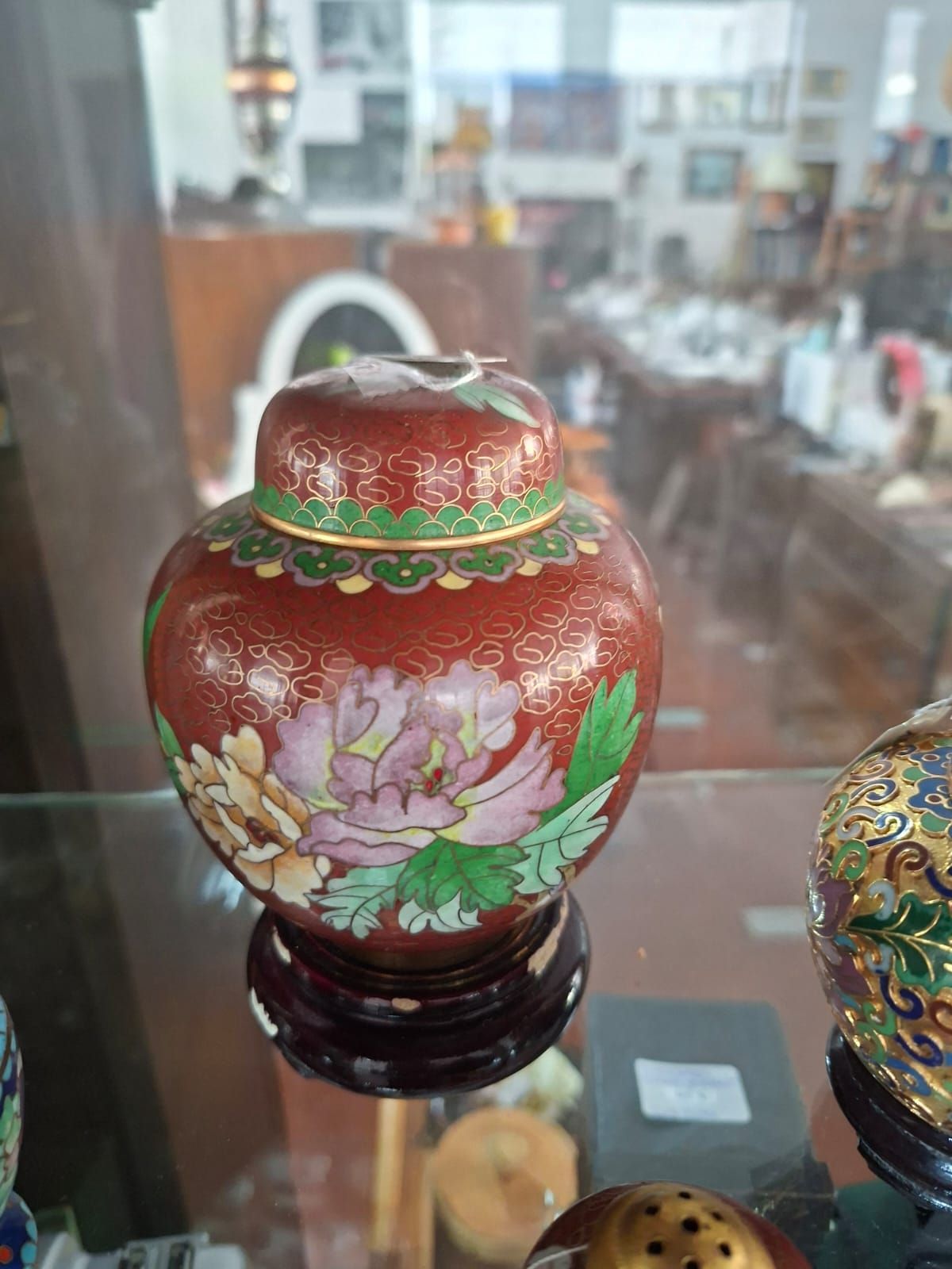 Un piccolo vaso rotondo in cloisonné rosso con motivo floreale di peonie e coperchio coordinato, esposto su un supporto di legno.