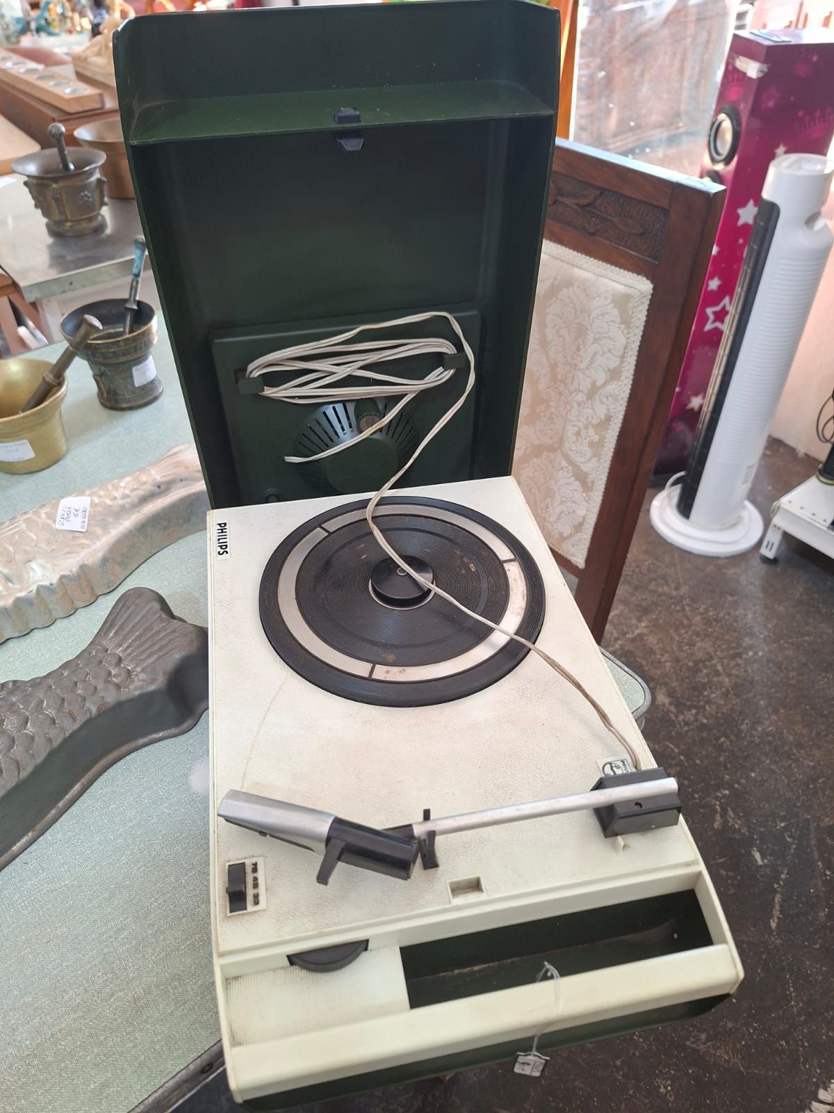 Un giradischi portatile vintage color crema con coperchio verde scuro, appoggiato su un tavolo all'interno di un negozio.