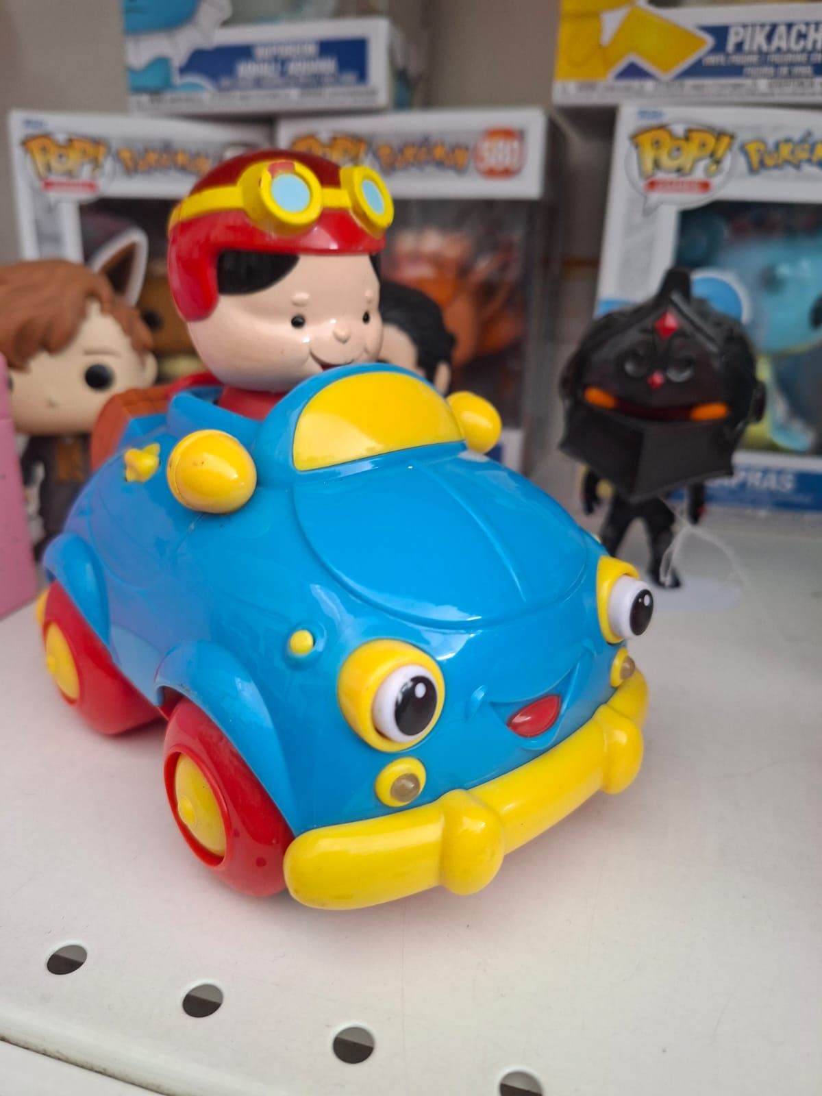 Un'auto giocattolo blu con finiture gialle e un personaggio dei cartoni animati sorridente al posto di guida, appoggiata su uno scaffale insieme a delle scatole di giocattoli.