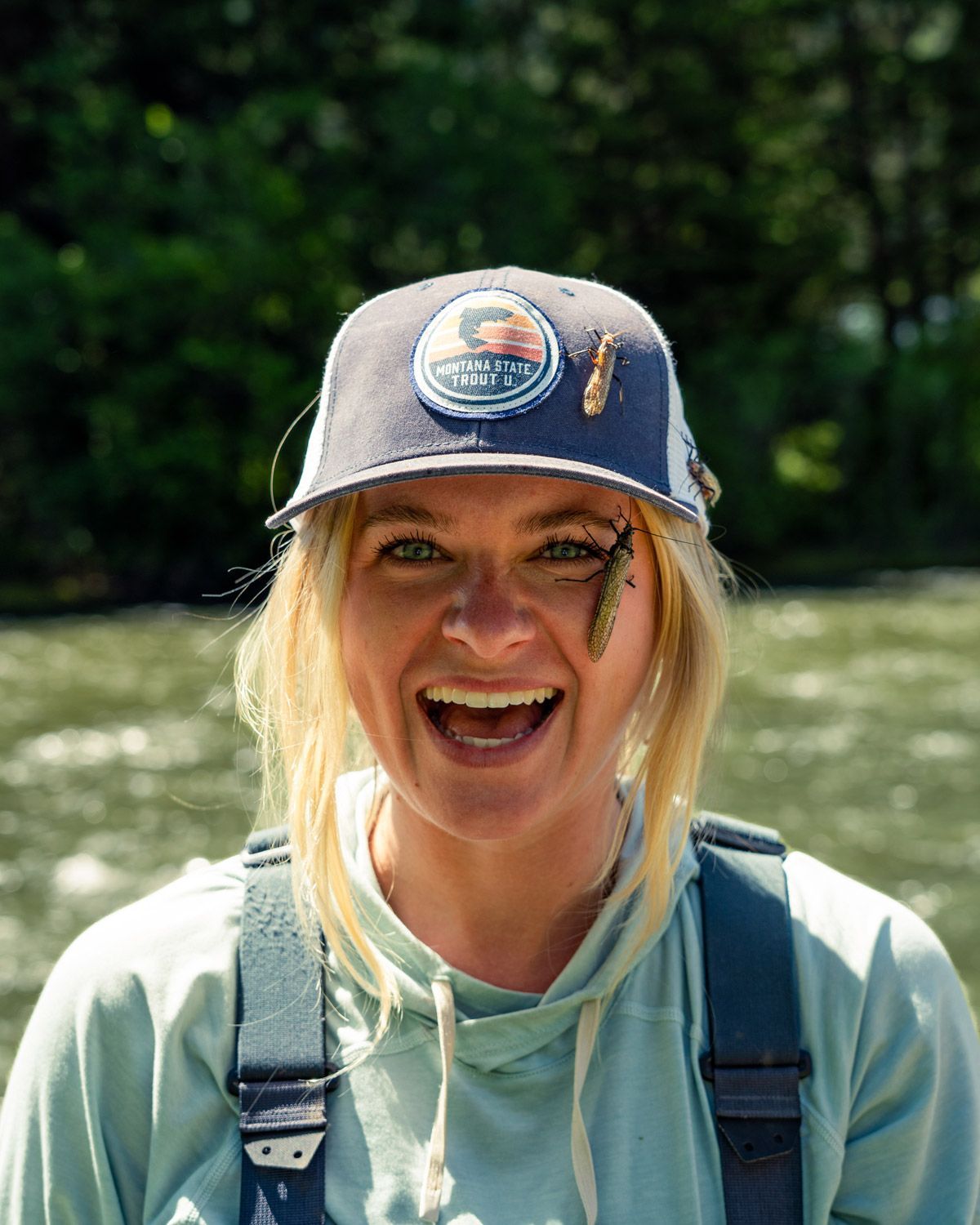 A woman wearing a hat and overalls is smiling in front of a river.