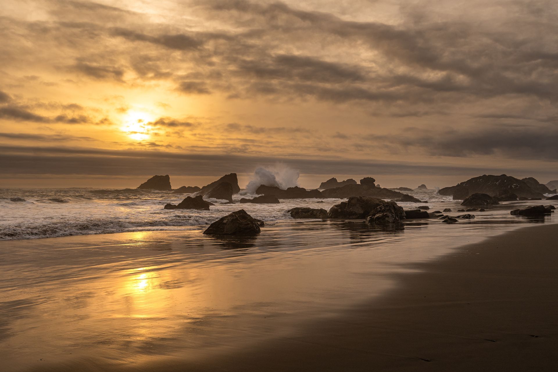 The sun is setting over the ocean and the waves are crashing on the rocks on the beach.