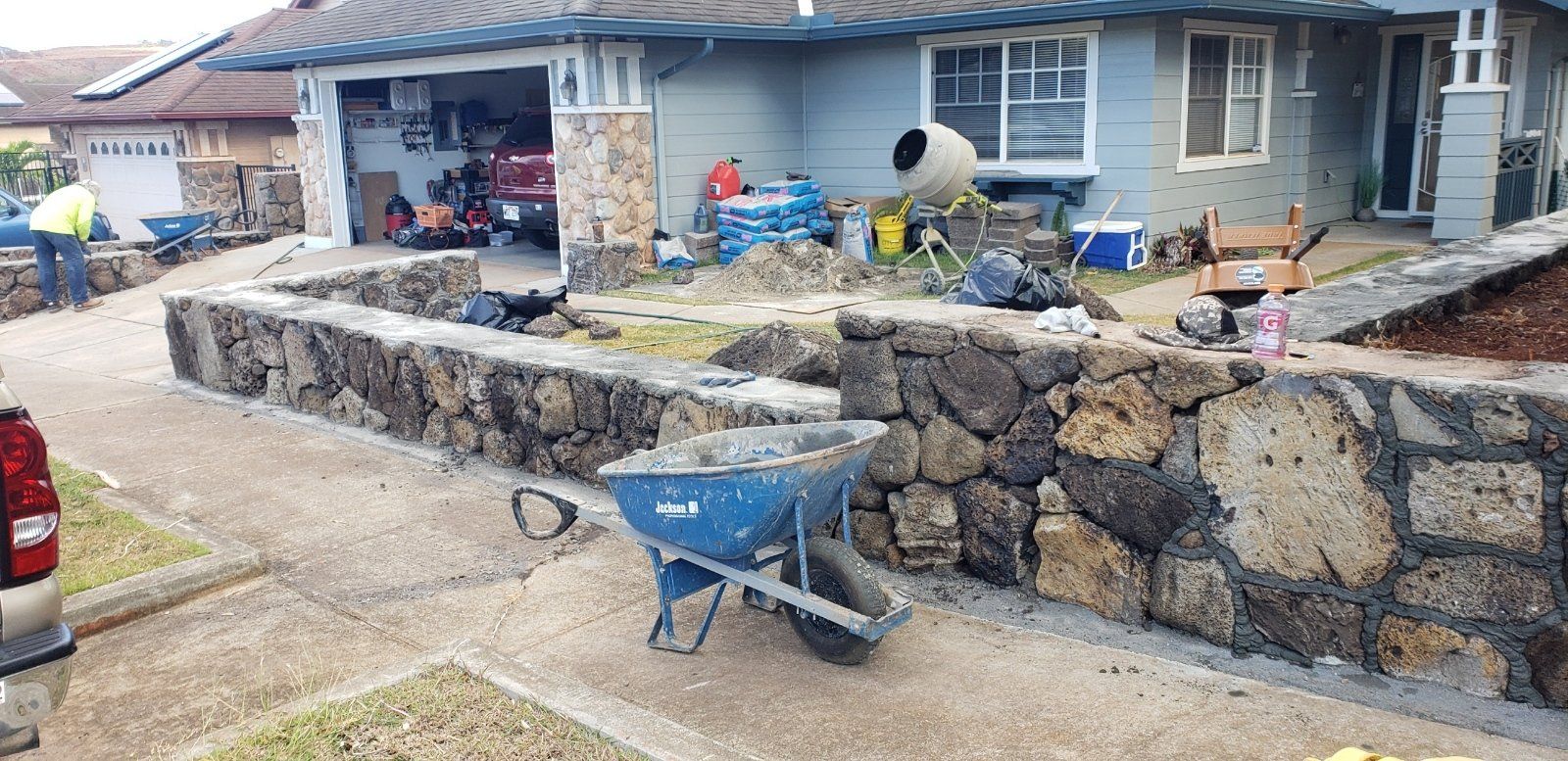 Front yard in Oahu with a stone retaining wall, blue wheelbarrow, and house under renovation debris