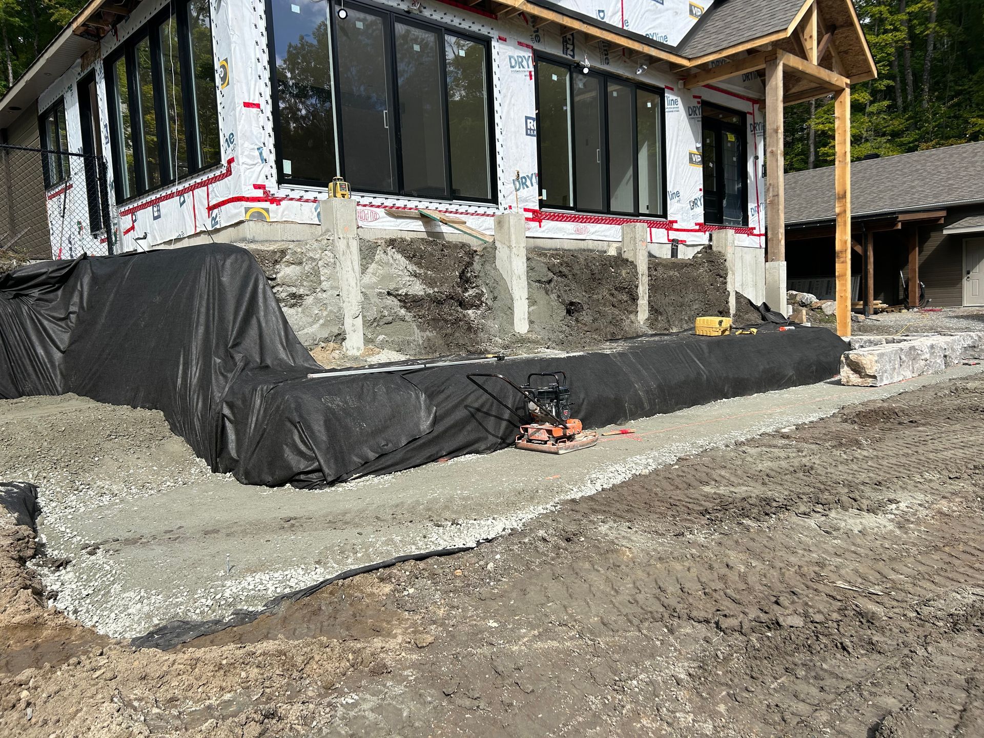 Vue extérieure d'une maison en construction avec un chemin en béton et une toile de paillage noire à sa base.
