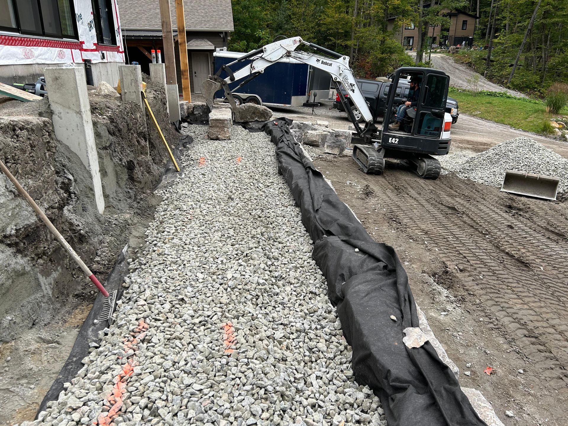 Chantier : caniveau de drainage en gravier recouvert de géotextile noir, excavatrice, bâtiment en construction.