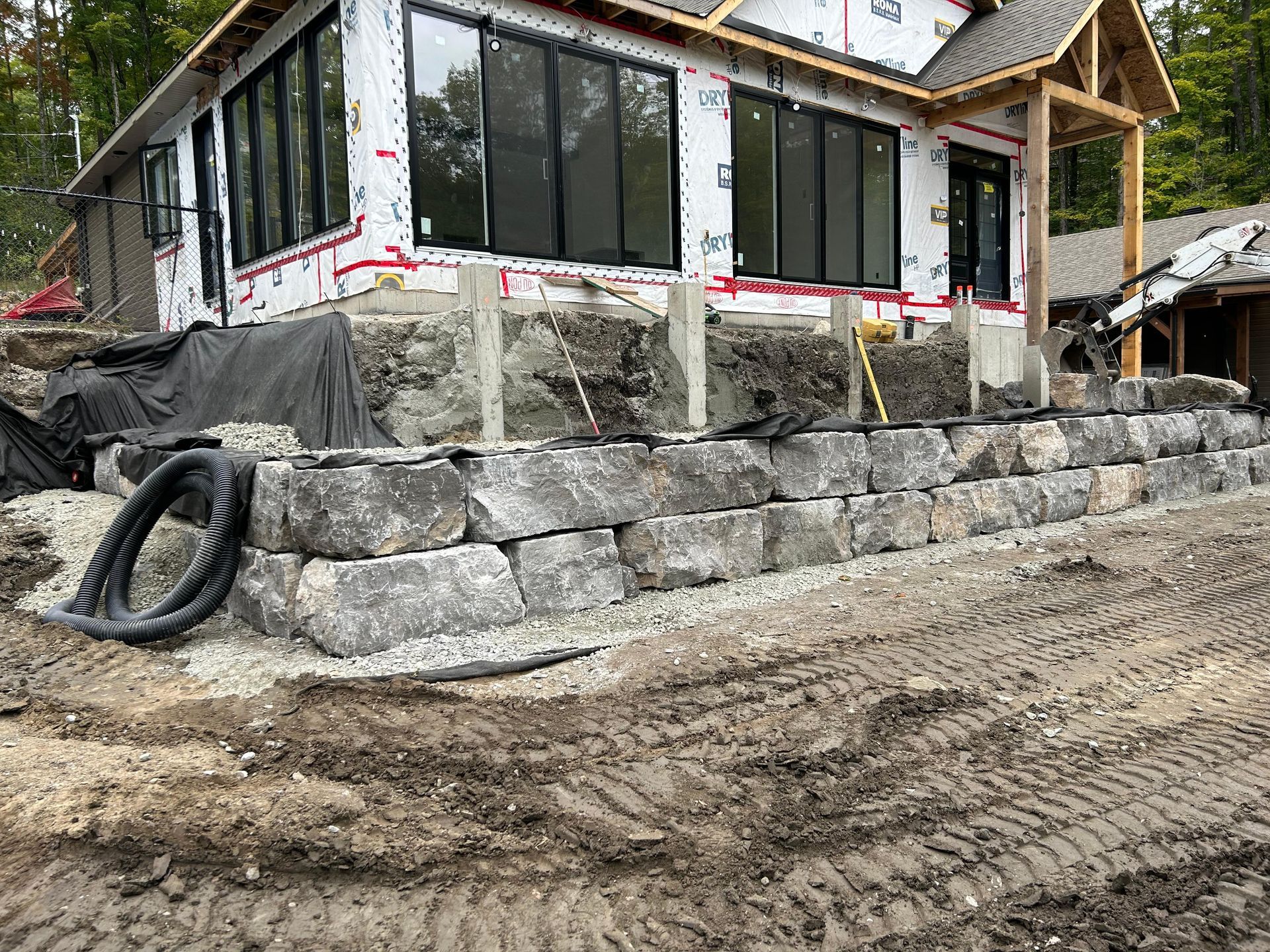 Mur de soutènement en pierre en construction devant une maison avec de grandes fenêtres.