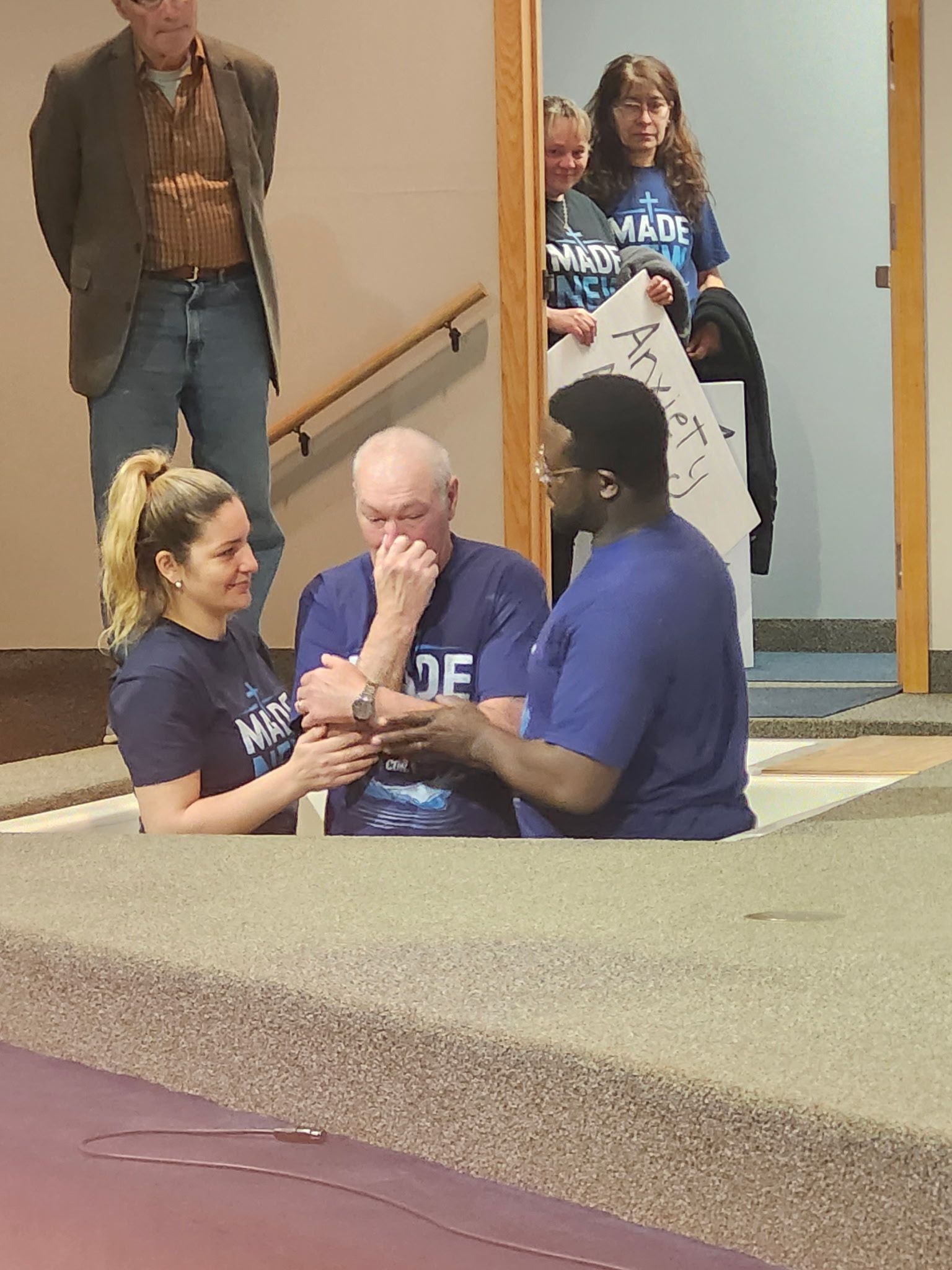 Three people in blue shirts kneel together on a gym floor during a ceremony or huddle.