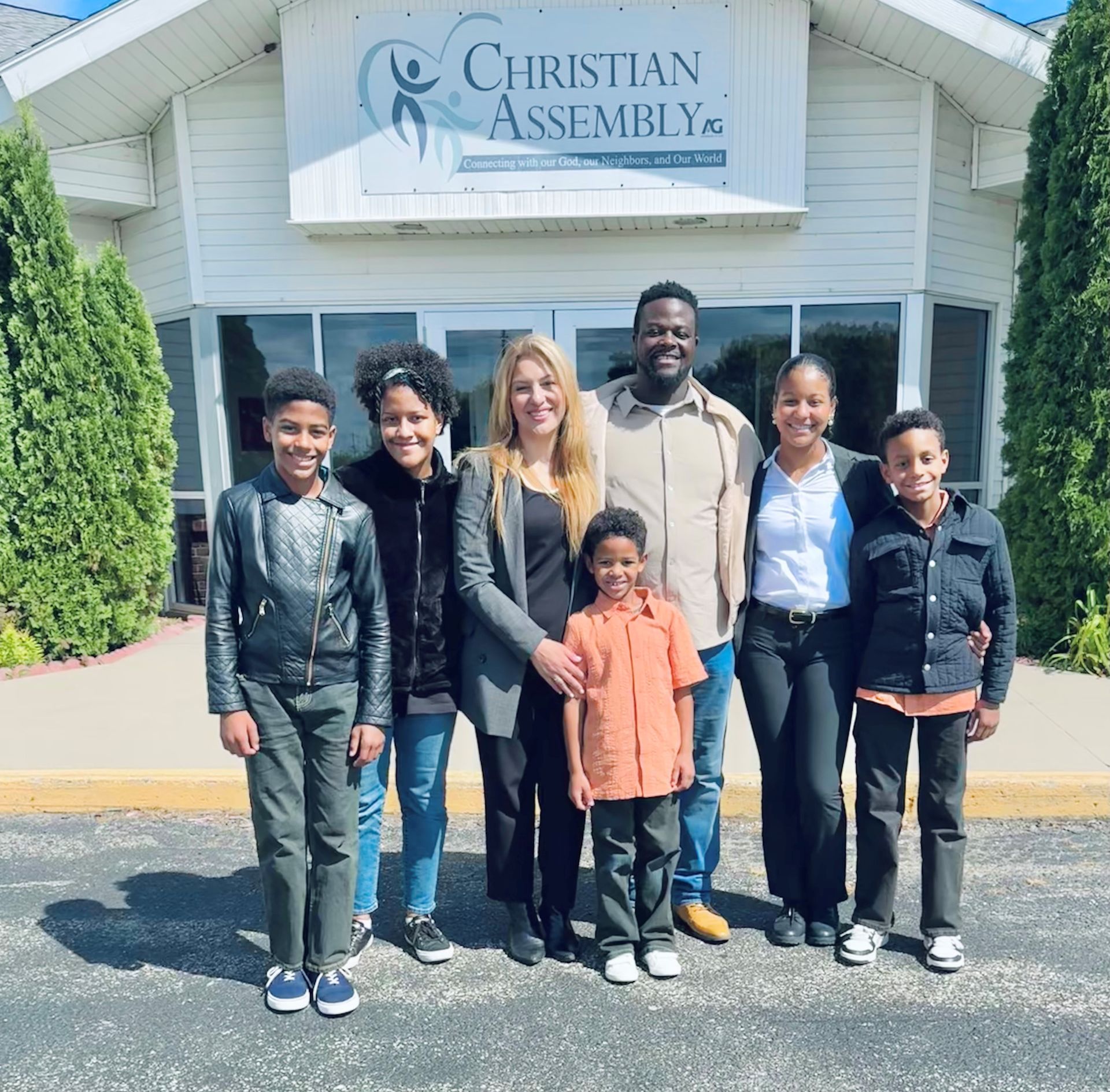 Group stands smiling outside Christian Assembly church sign.