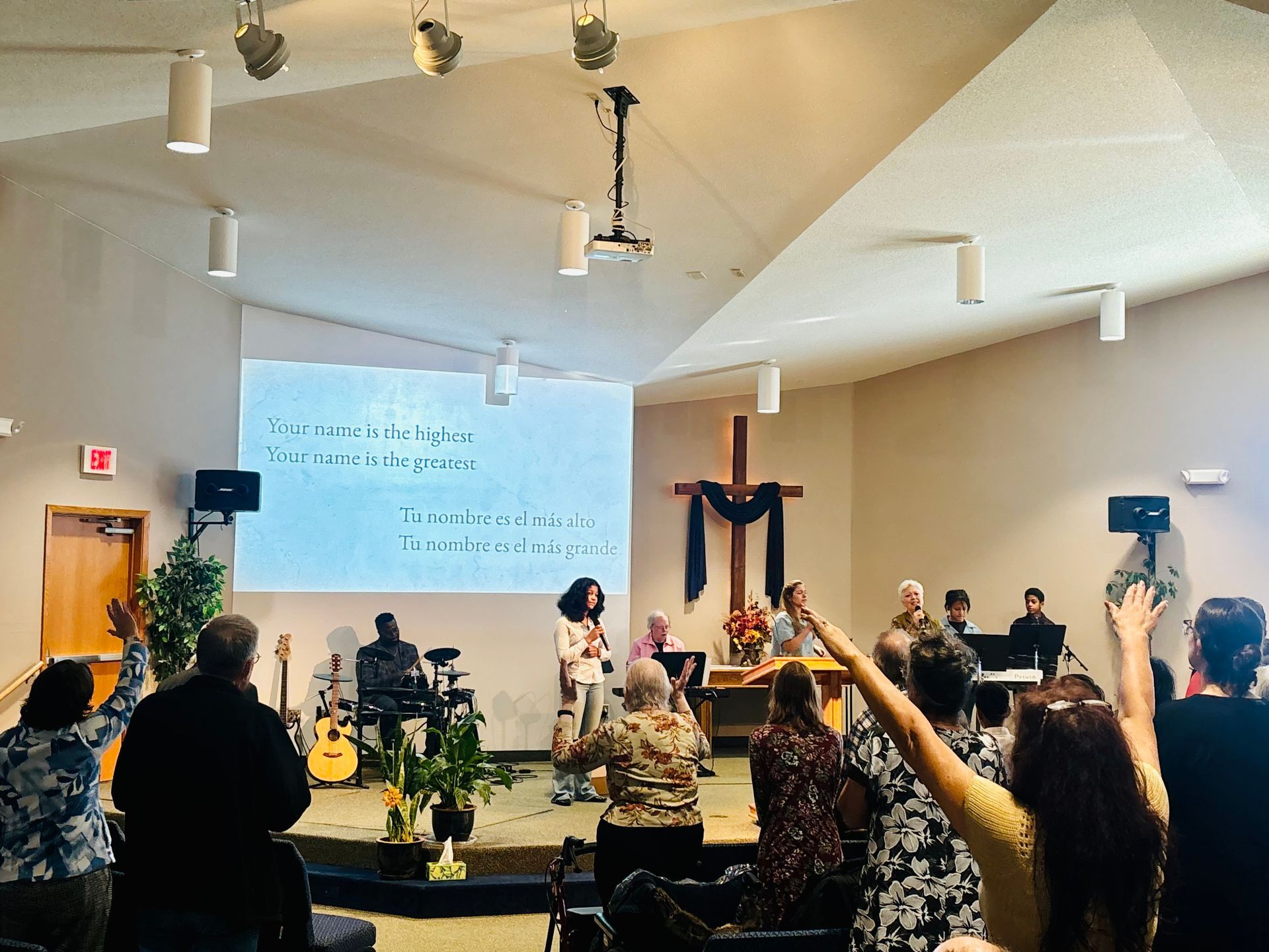 Audience in church hall facing a speaker at the front, with raised hands and projected lyrics on the wall.