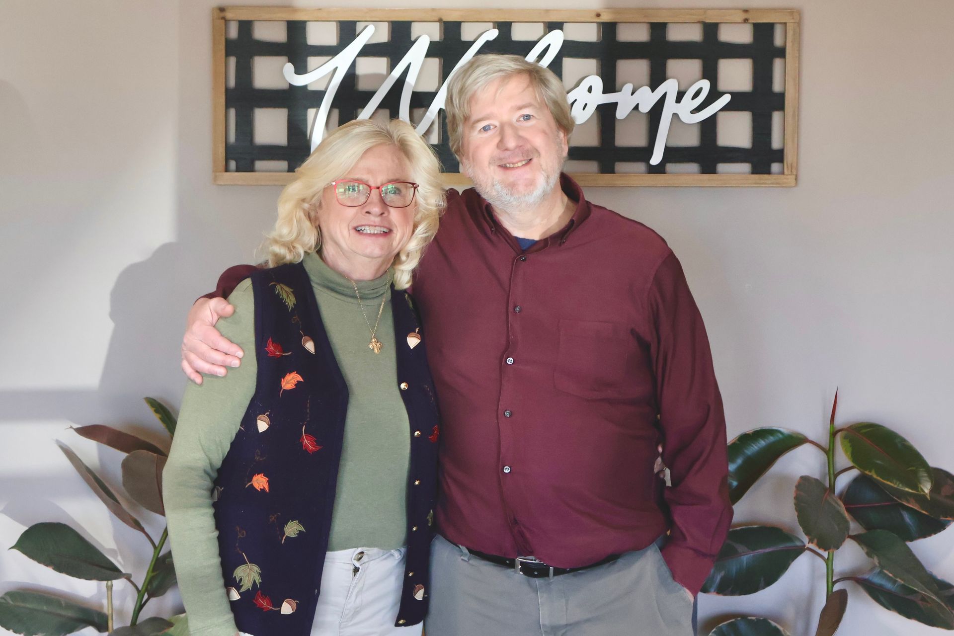 Two smiling people standing together indoors in front of a welcome sign and plants.