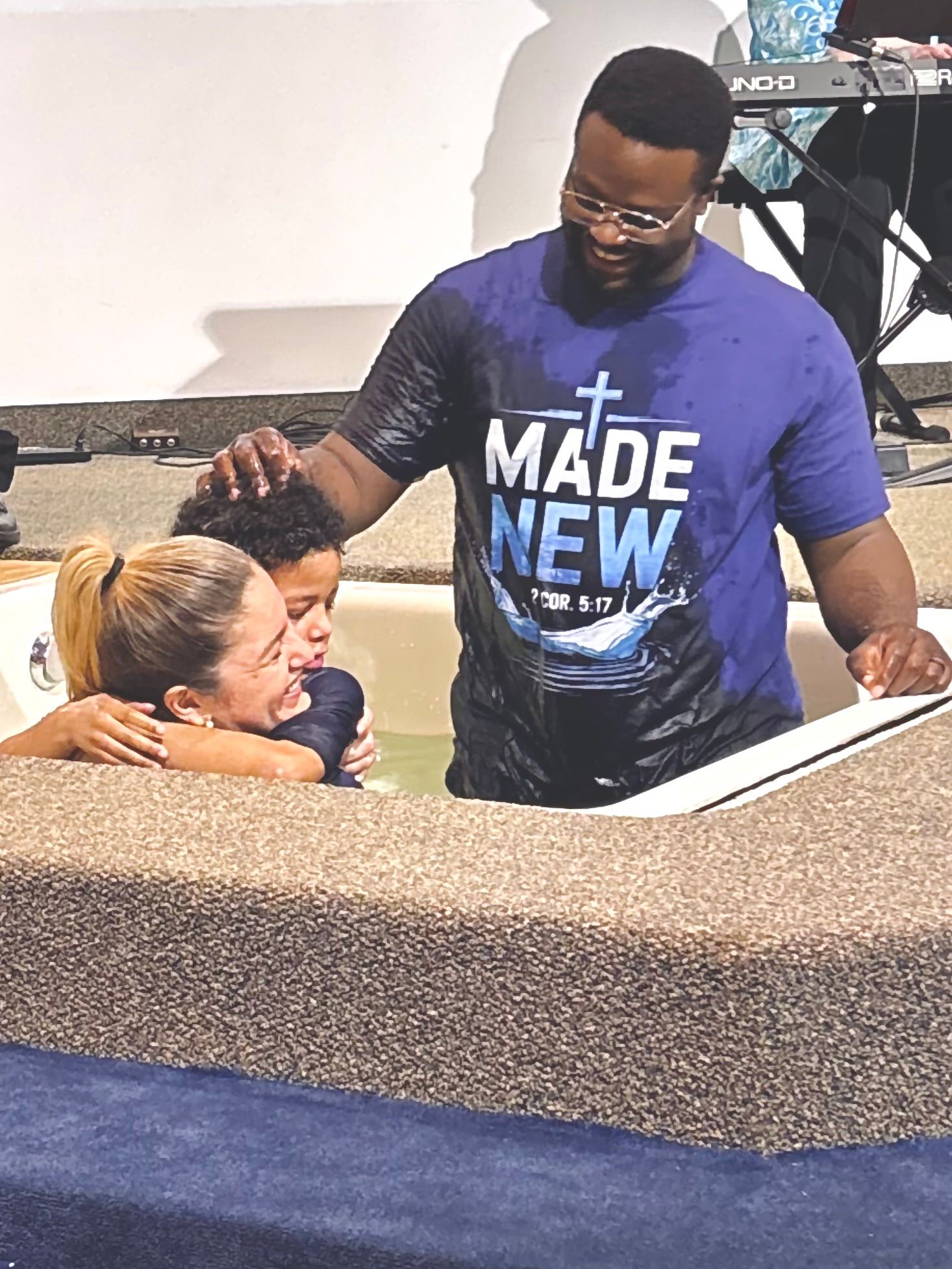 Man in a blue “Made New” shirt praying with a person seated in a church baptistry