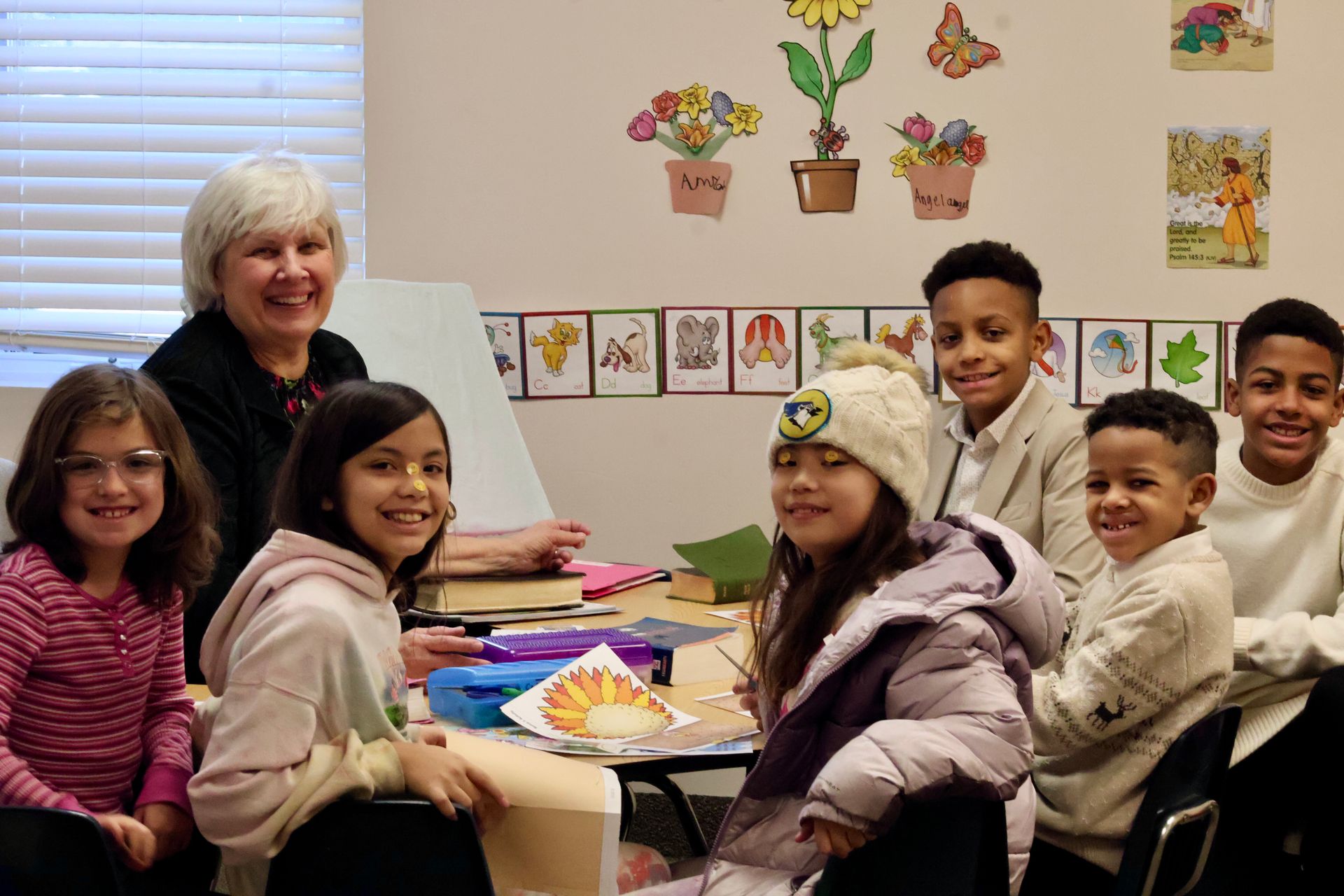 Teacher and children sit around a classroom table, smiling during an art activity.