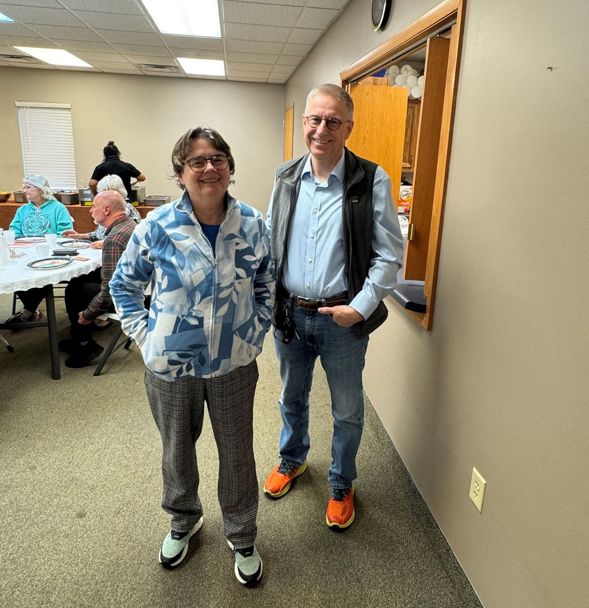 Two people standing in a hallway, smiling beside a bulletin board, with tables and people in the background.
