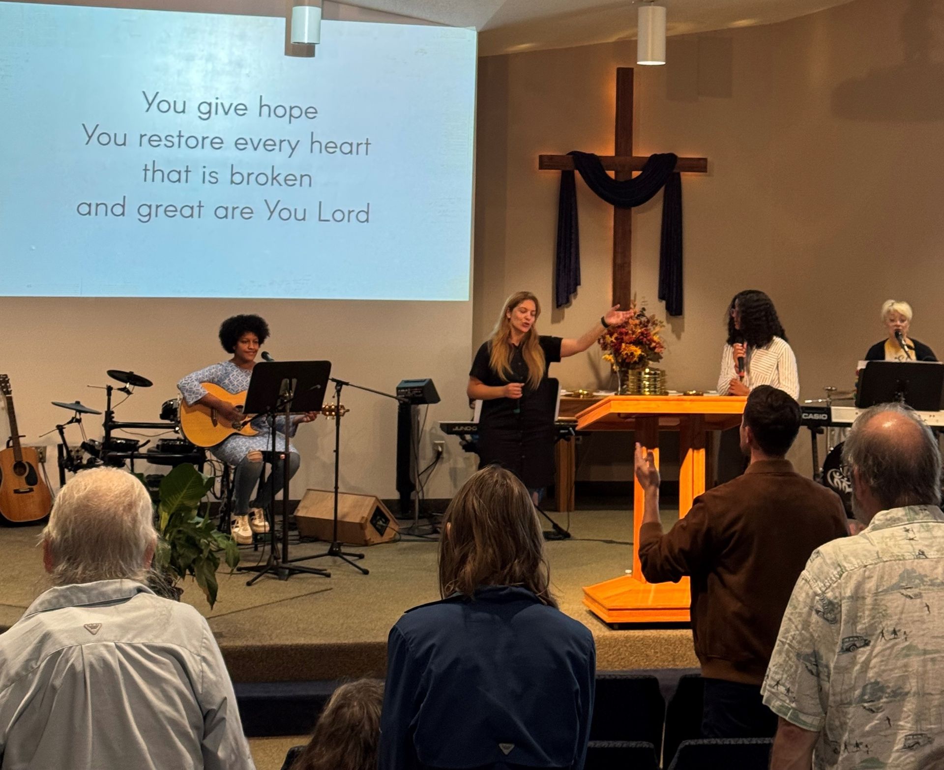 Church worship service with musicians, speaker at lectern, cross on wall, and seated congregation