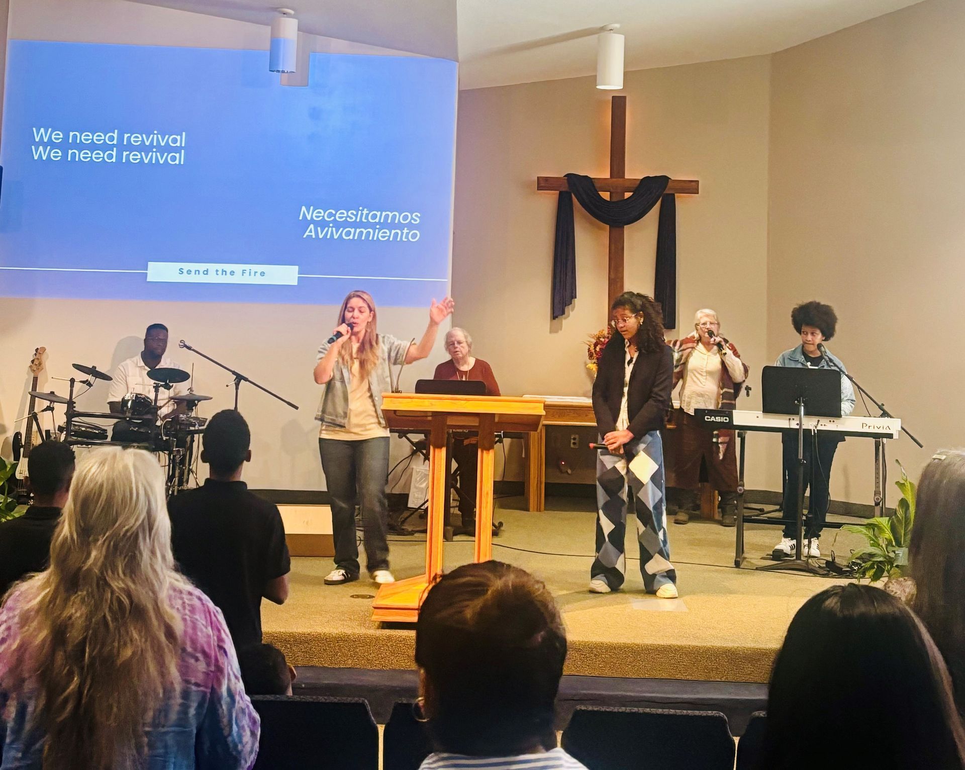 Worship band on church stage with singers, keyboardist, drums, and a large cross behind them