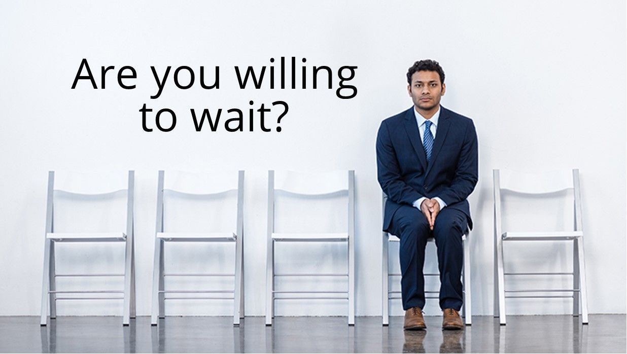 A man in a suit sits on one of five empty chairs against a white wall with the text