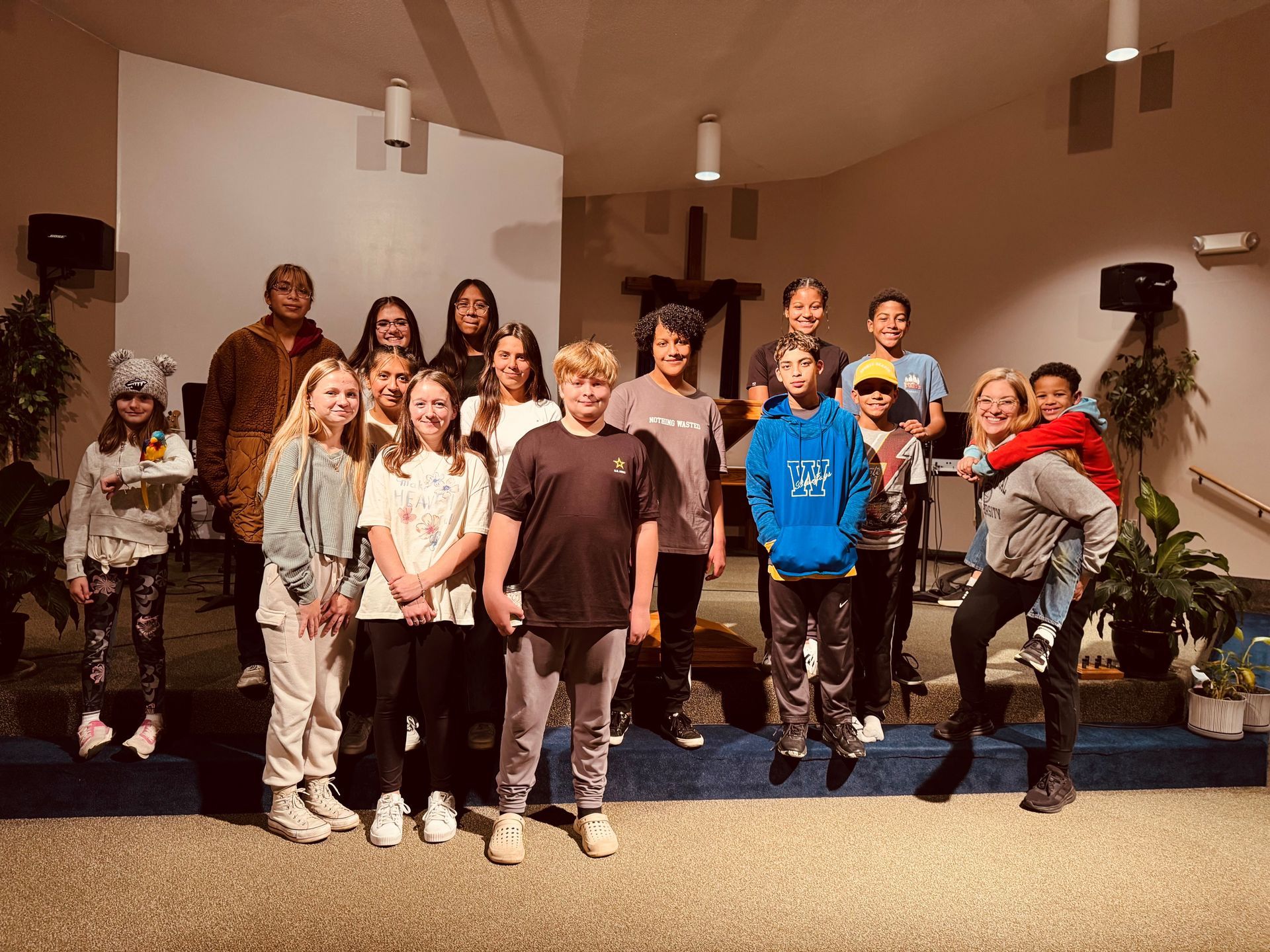 Group of children posing on a stage in a church, with potted plants and white walls behind them.