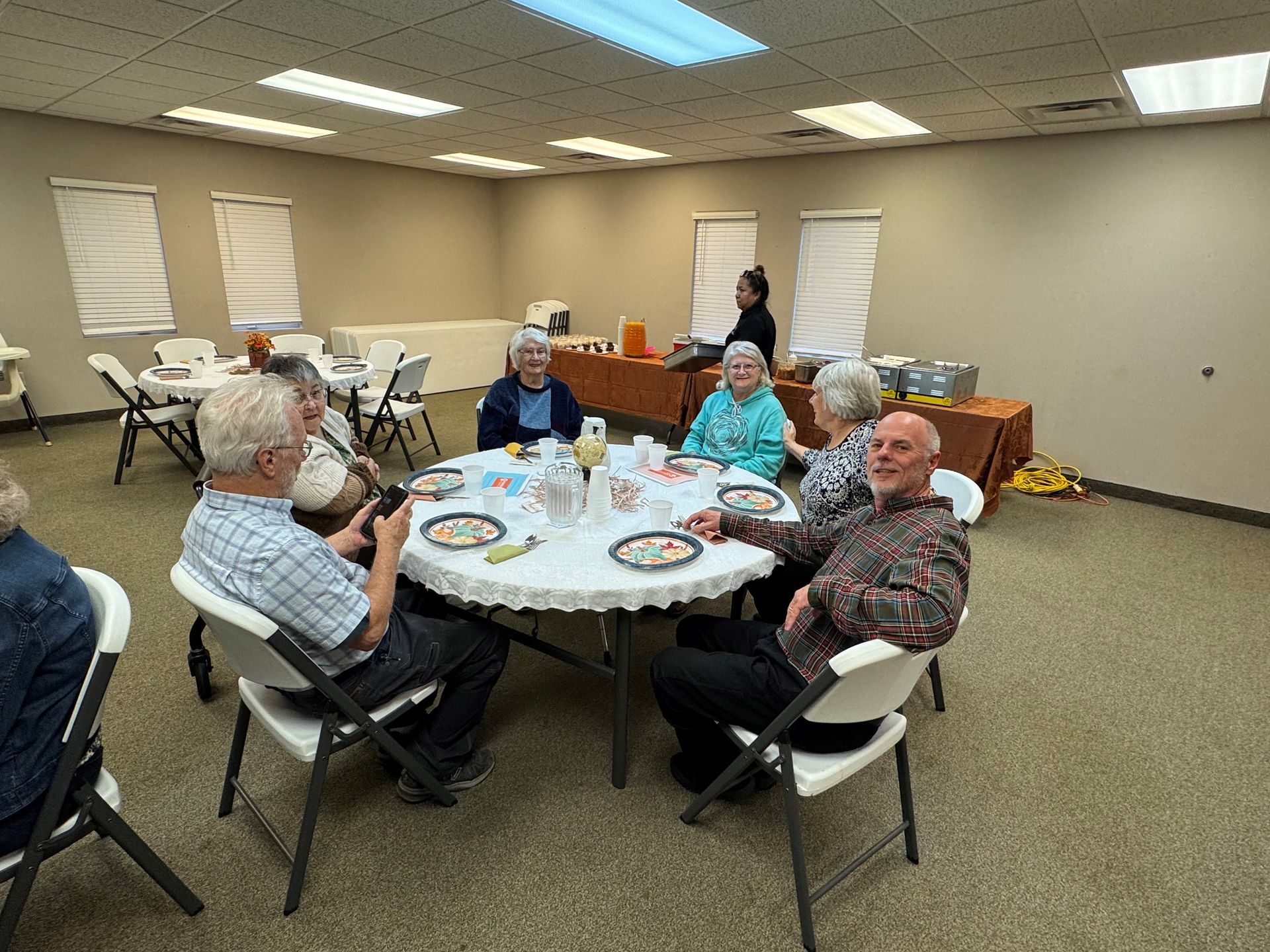 People seated around a round table in a community room, eating and talking with plates and drinks nearby.