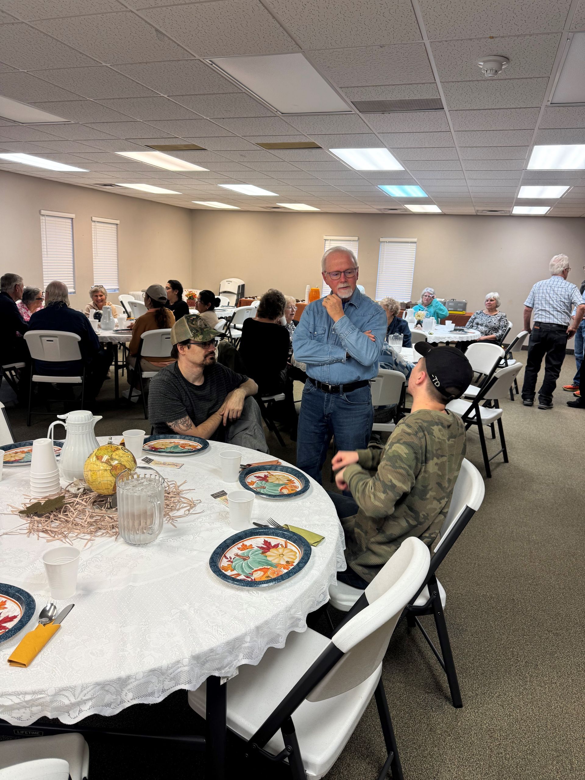 Community banquet in a hall, with people seated at round tables and one man standing and speaking.