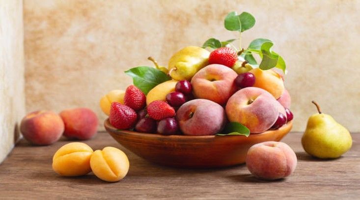 Assorted fresh fruit in a bowl on a wooden table, with peaches, apples, pears, and strawberries against a rustic wall