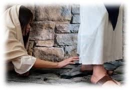 Person kneeling by a stone wall, reaching toward another person’s white robe and sandals